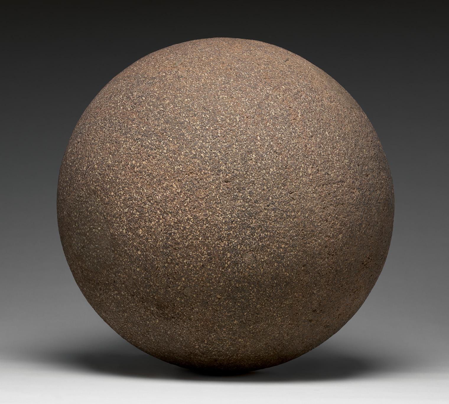 A large, weathered brown stone sphere rests on a gray surface, casting a soft shadow against a dark background
