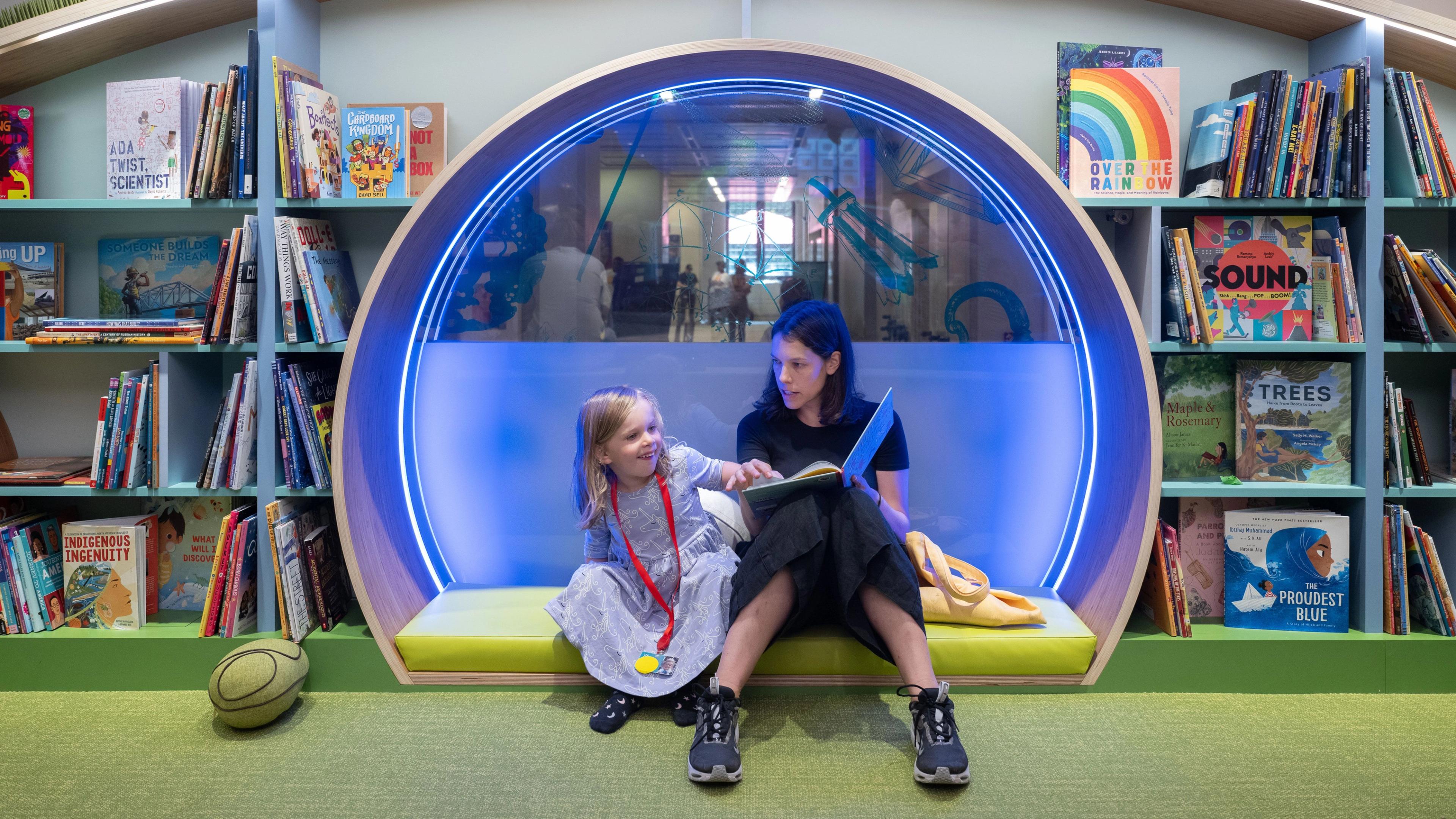 An adult and child sit inside a colorful reading nook located in between shelves of children's books.
