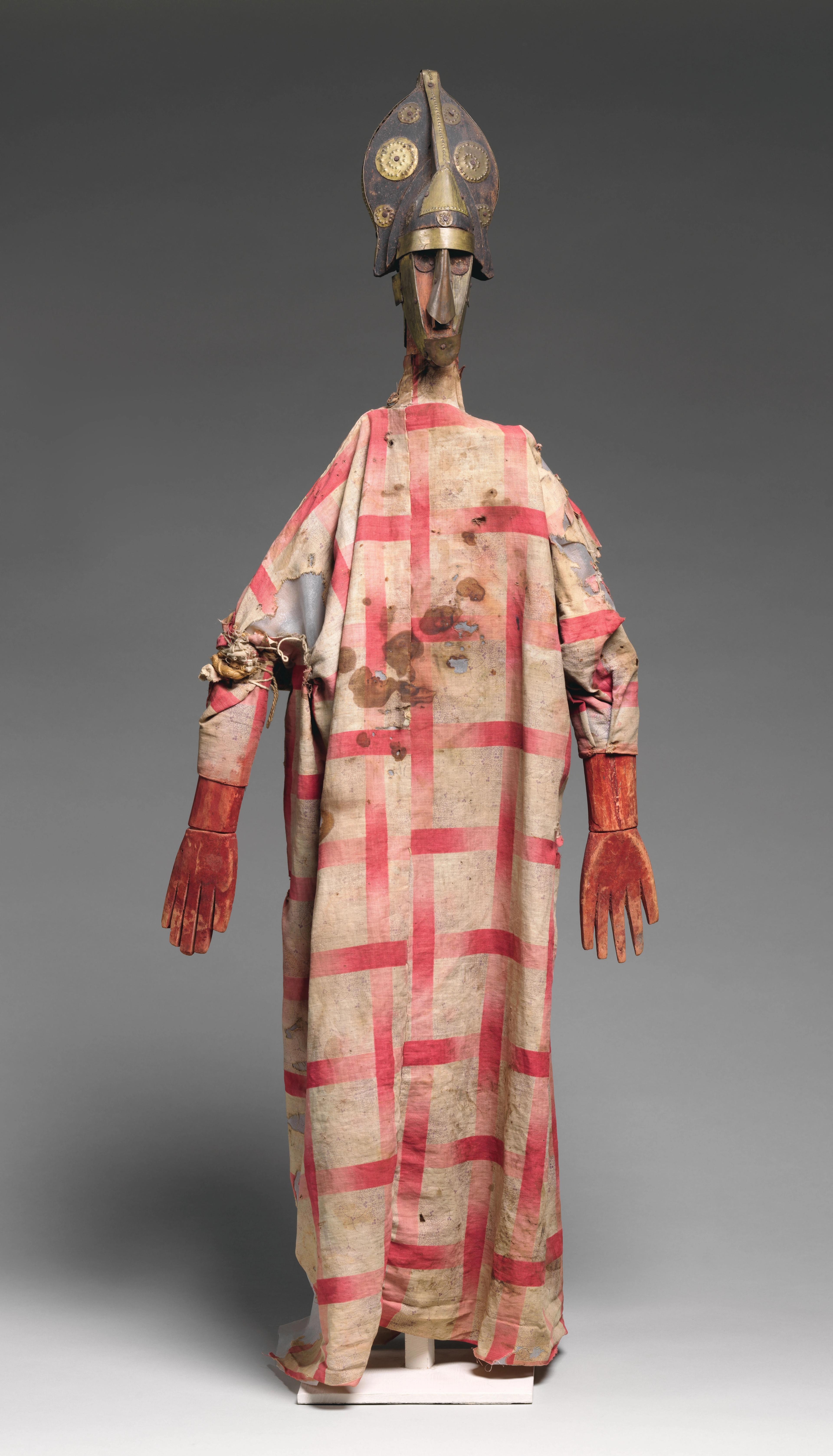 Carved wood marionette with a headdress wearing a red plaid gown and red gloves.