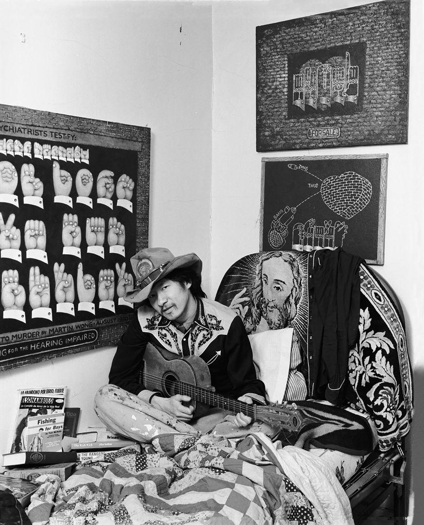 Black and white photograph of Martin Wong seated holding a guitar on a twin bed surrounded by his artworks hanging on the walls to either side of him.