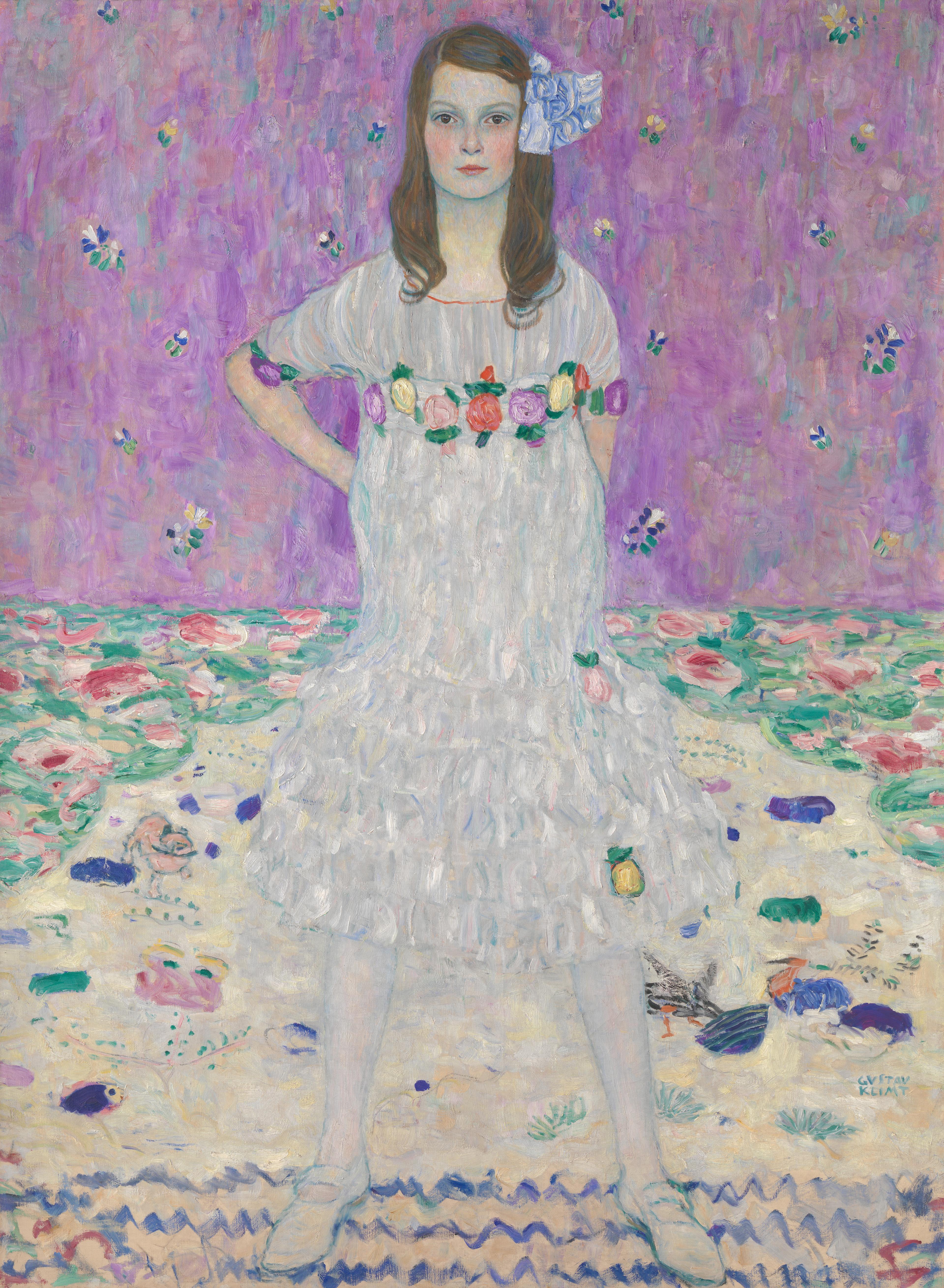A young girl poses with a stoic face and a hand on her hip. She stands on a carpet with whimsical designs, and her dress and hair are accented with flowers.