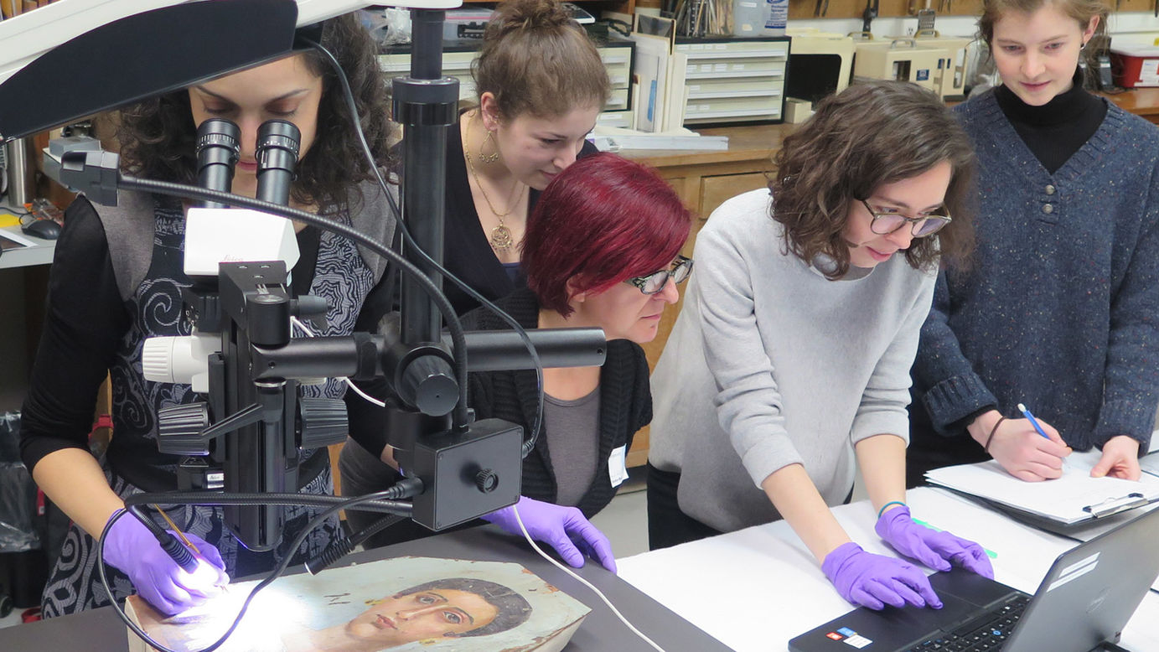 Five individuals working around a table at a laptop and using a microscope to look closely at an artwork.