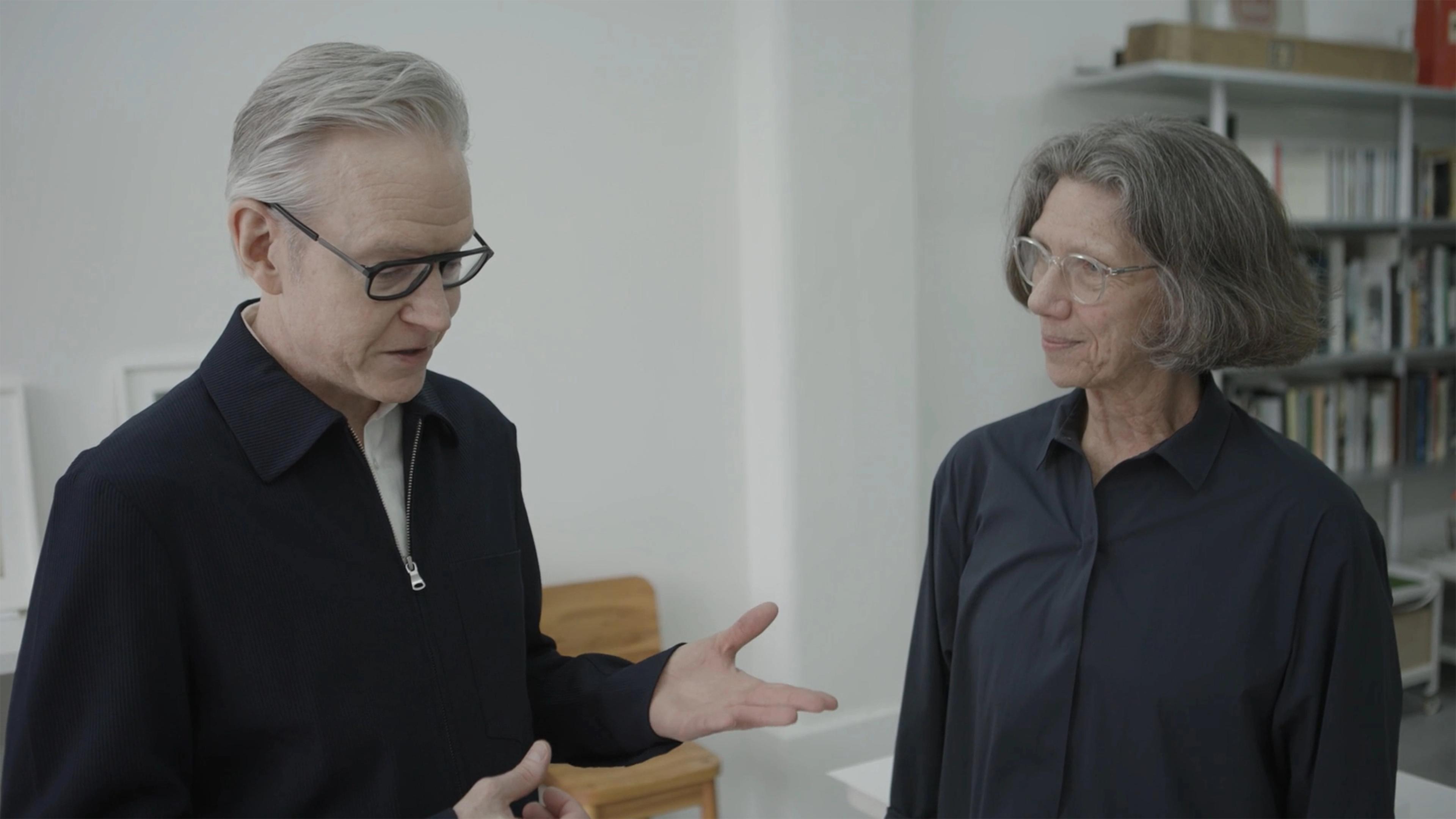Two people wearing dark shirts and glasses converse in a bright room, with one gesturing while the other listens.