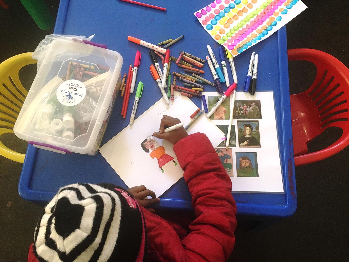 Ponele, seated at a blue table, draws a portrait of her mother.