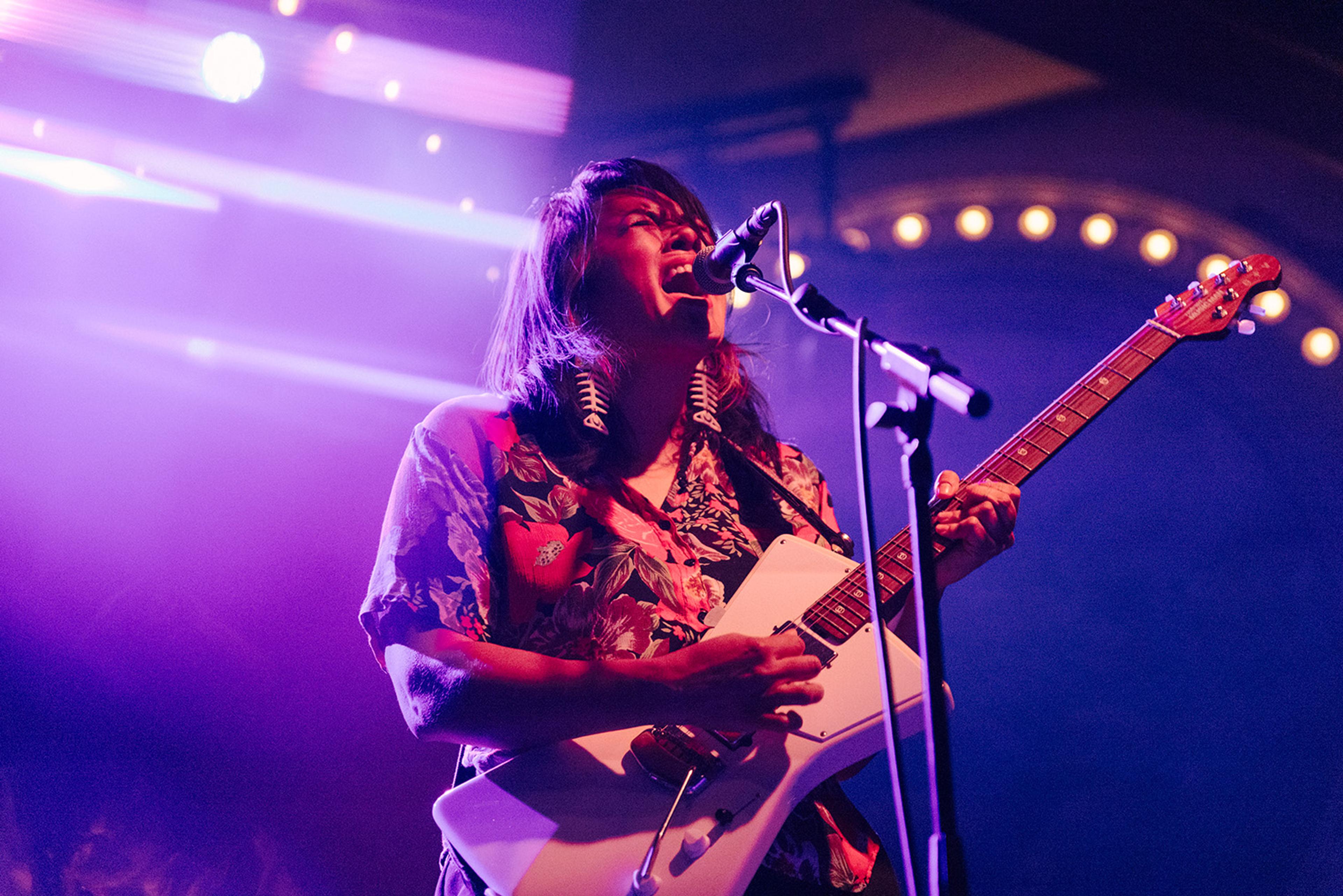 A long-haired musician sings into a microphone while playing a white electric guitar onstage, bathed in vivid purple light and warm spotlights.