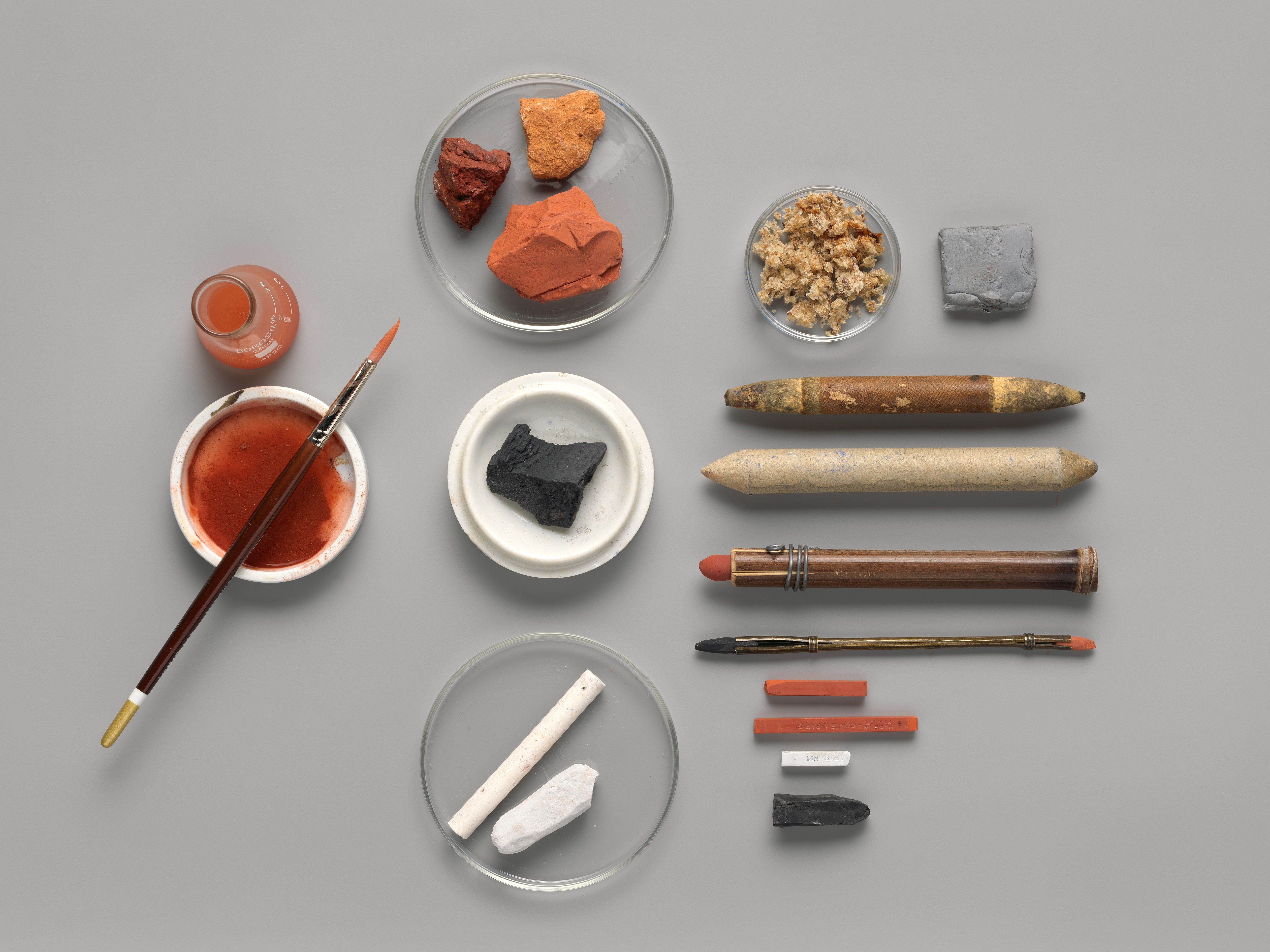 An artful arrangement of specialized tools for drawing with chalk, including multiple sticks of chalks, chalk-holders, and a small cup of water and a brush.