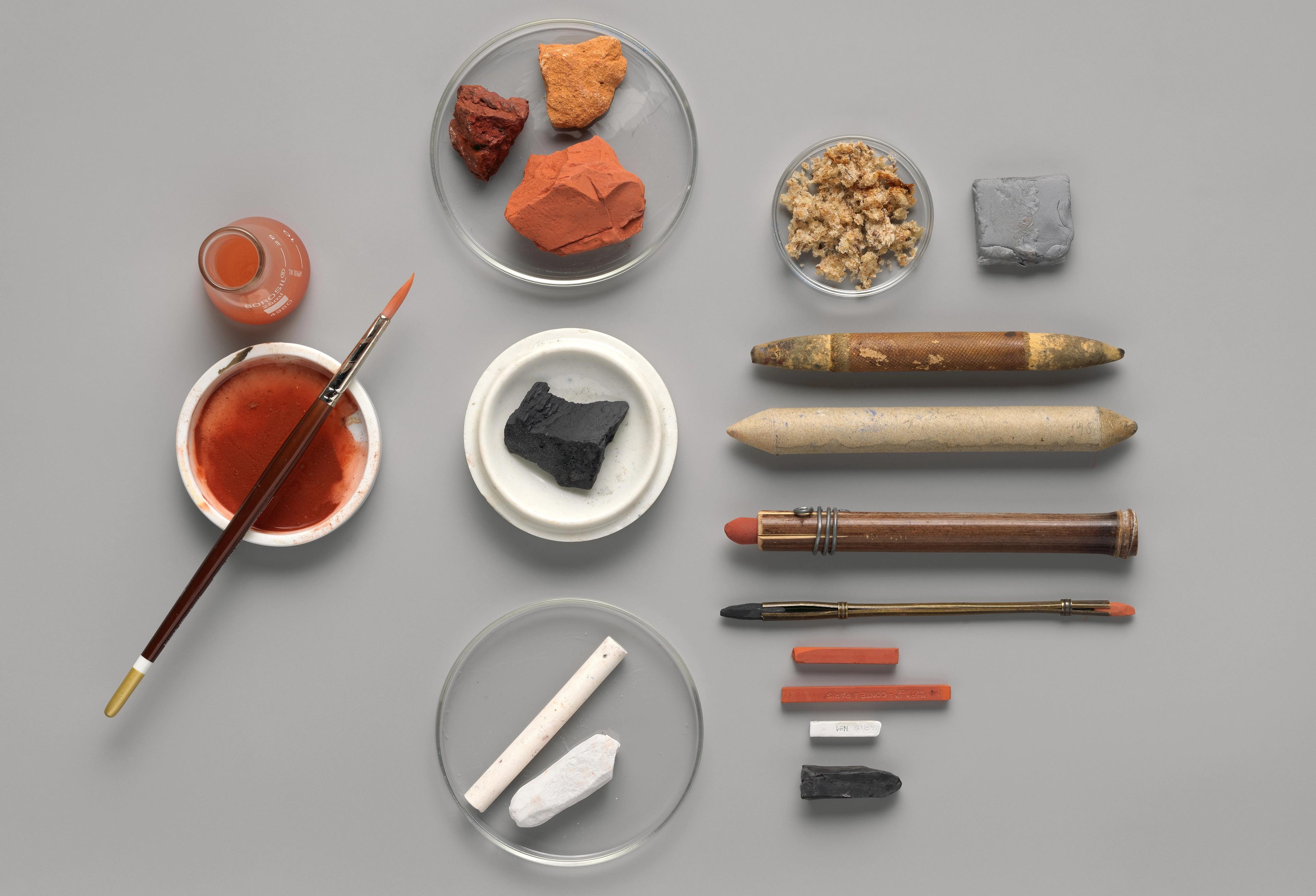 An artful arrangement of specialized tools for drawing with chalk, including multiple sticks of chalks, chalk-holders, and a small cup of water and a brush.