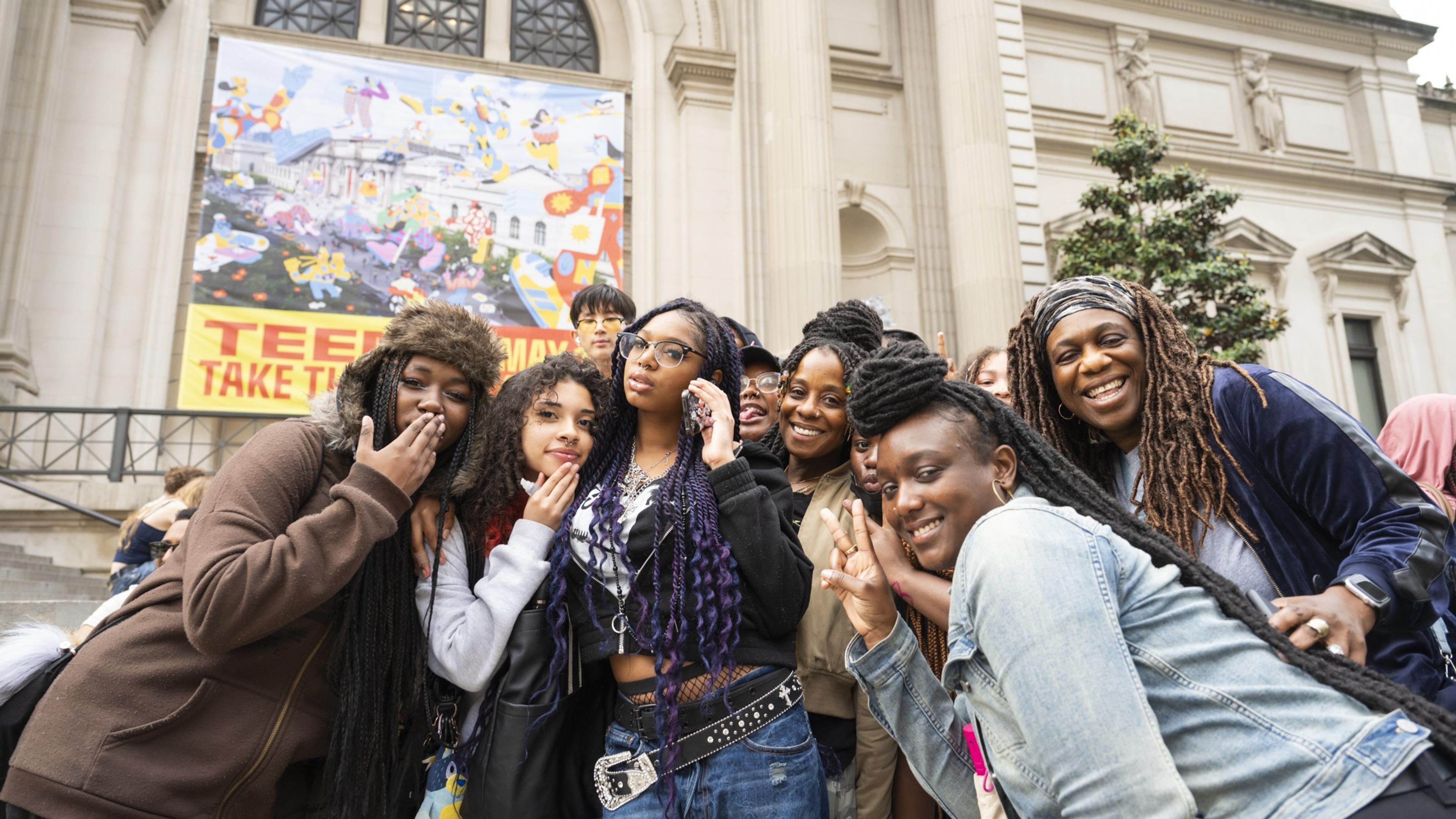 A group of teens gathering in front of a Museum with a poster that says "Teens Take The Met!"
