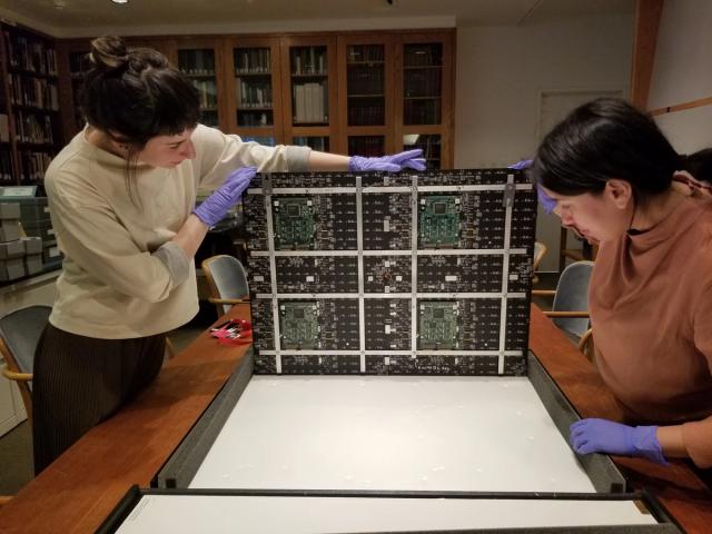 Two people wearing gloves examine a large electronic circuit board laid out on a table in a room lined with bookshelves.