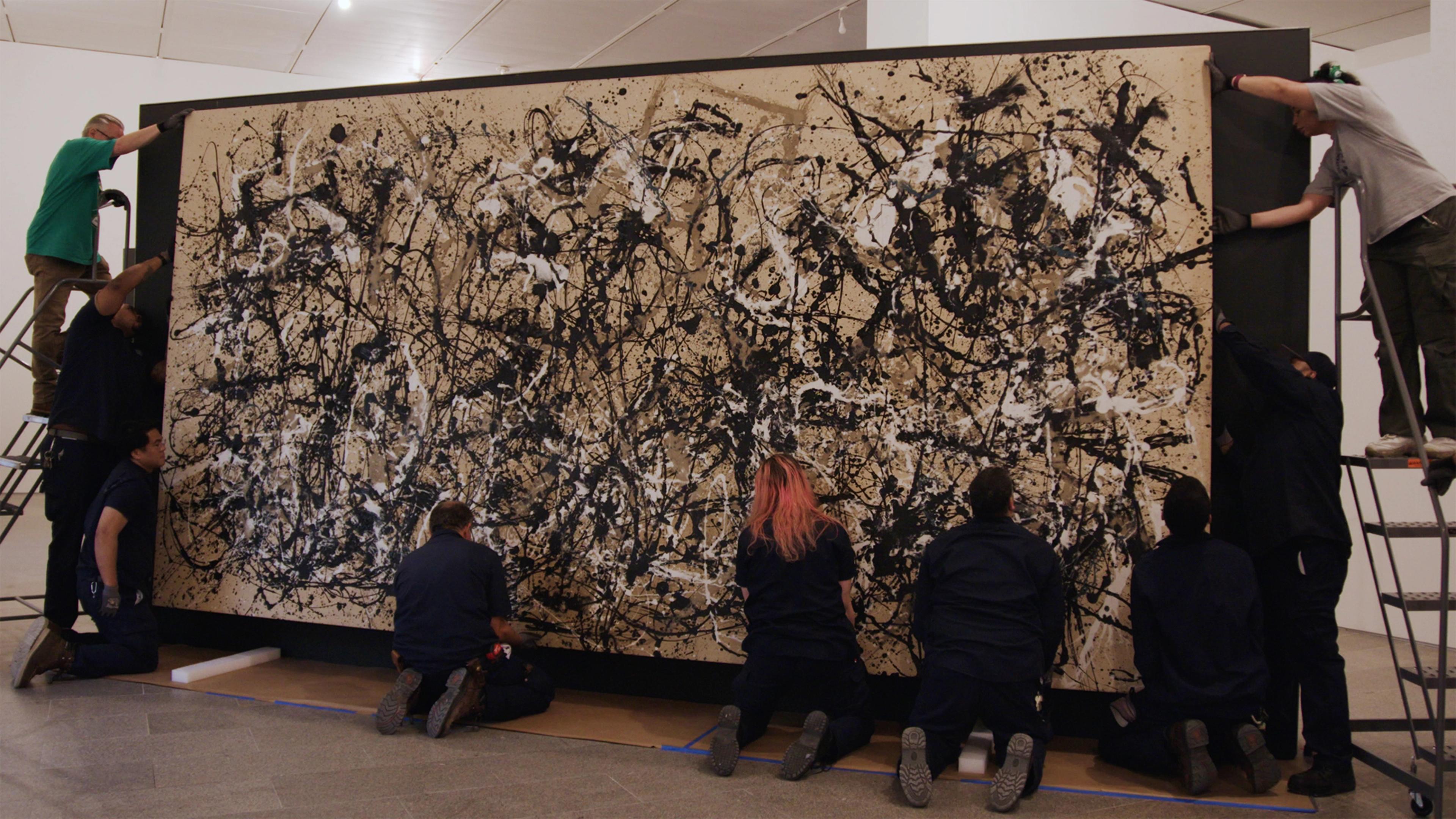 Several people install a monumental black-and-beige abstract splatter painting in a gallery, working together with care and concentration.