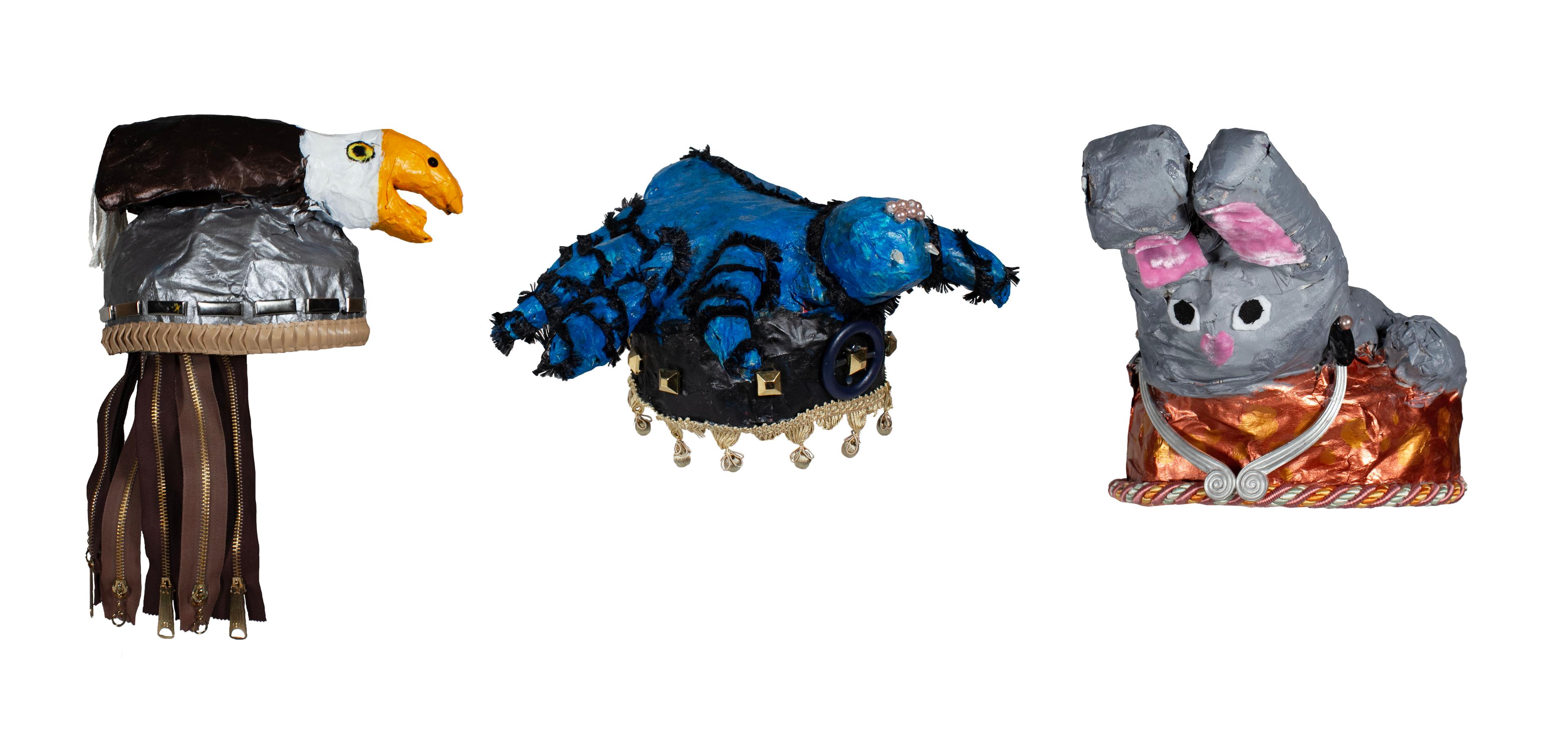 Sculptures of three animals represented as helmets created with recycled material, cardboard, acrylic, and papier mâché. A sculpture of a brown bald eagle with white face and yellow beak sits atop a shiny silver-colored helmet with several long, brown tassels hanging from the back composed of clothing zippers. A sculpture of a blue spider with black segmented joints sits atop a dark blue helmet with small beige accents hanging from the lower hat brim. A sculpture of a gray bunny with pink nose and pink inner ears sits atop a shiny copper-colored helmet with silver-colored ornamental trim.