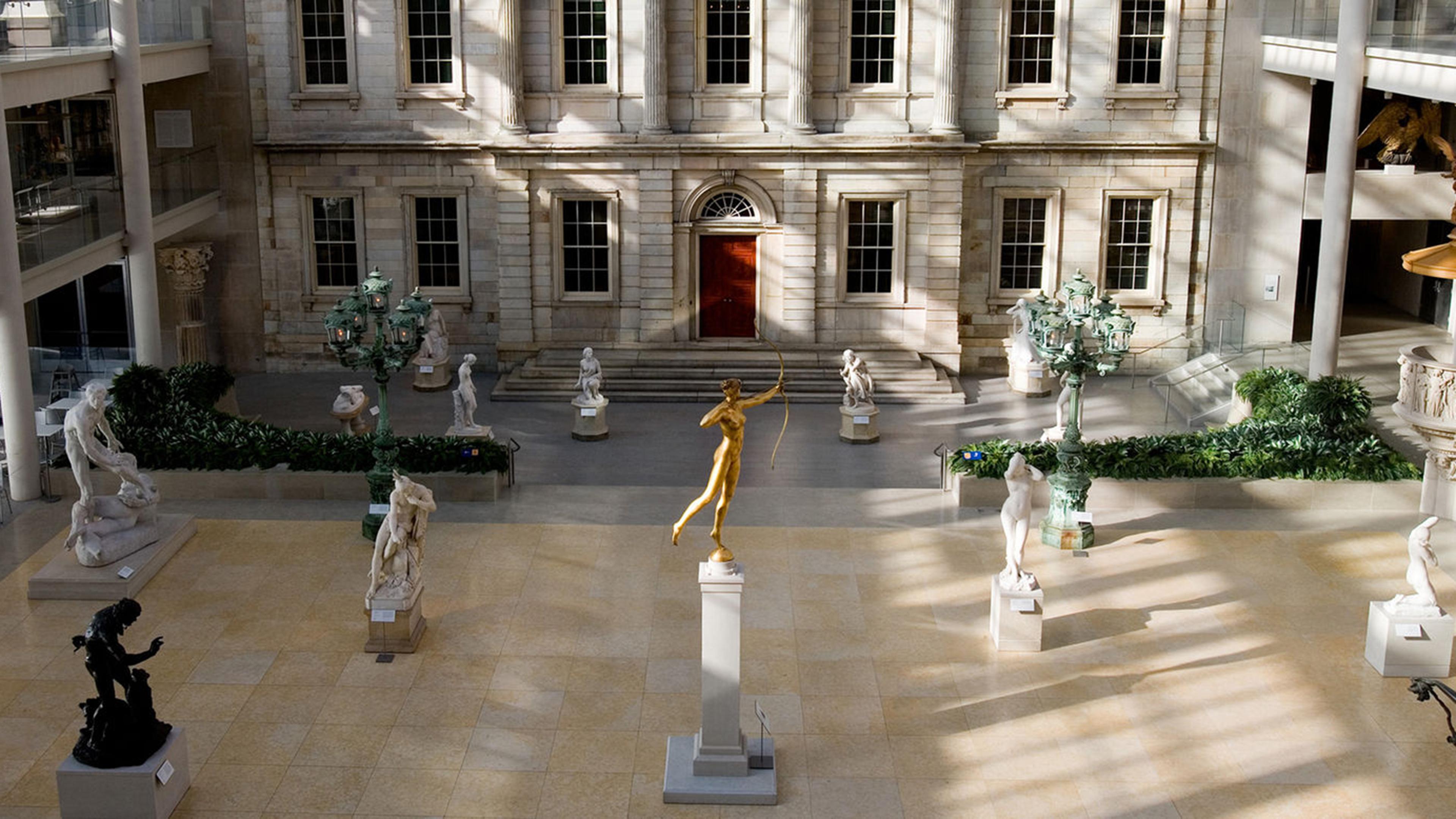 Large indoor courtyard with classical statues, including a gold figure on a pedestal, under a high glass ceiling. Sunlight casts shadows on the floor.