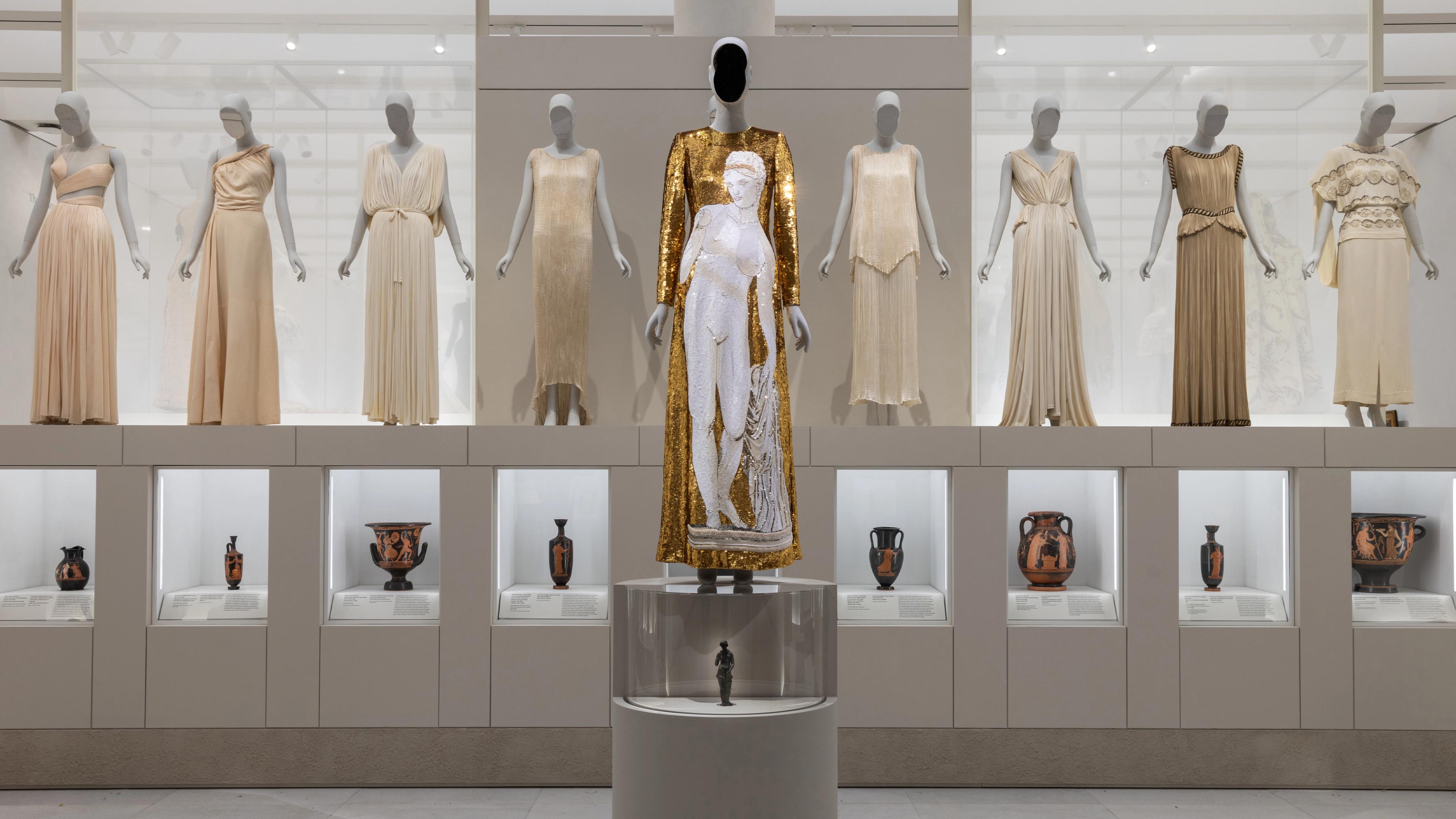 A gallery display features mannequins dressed in elegant gowns, with a central mannequin in a gold and white outfit, surrounded by ancient pottery in glass cases below.