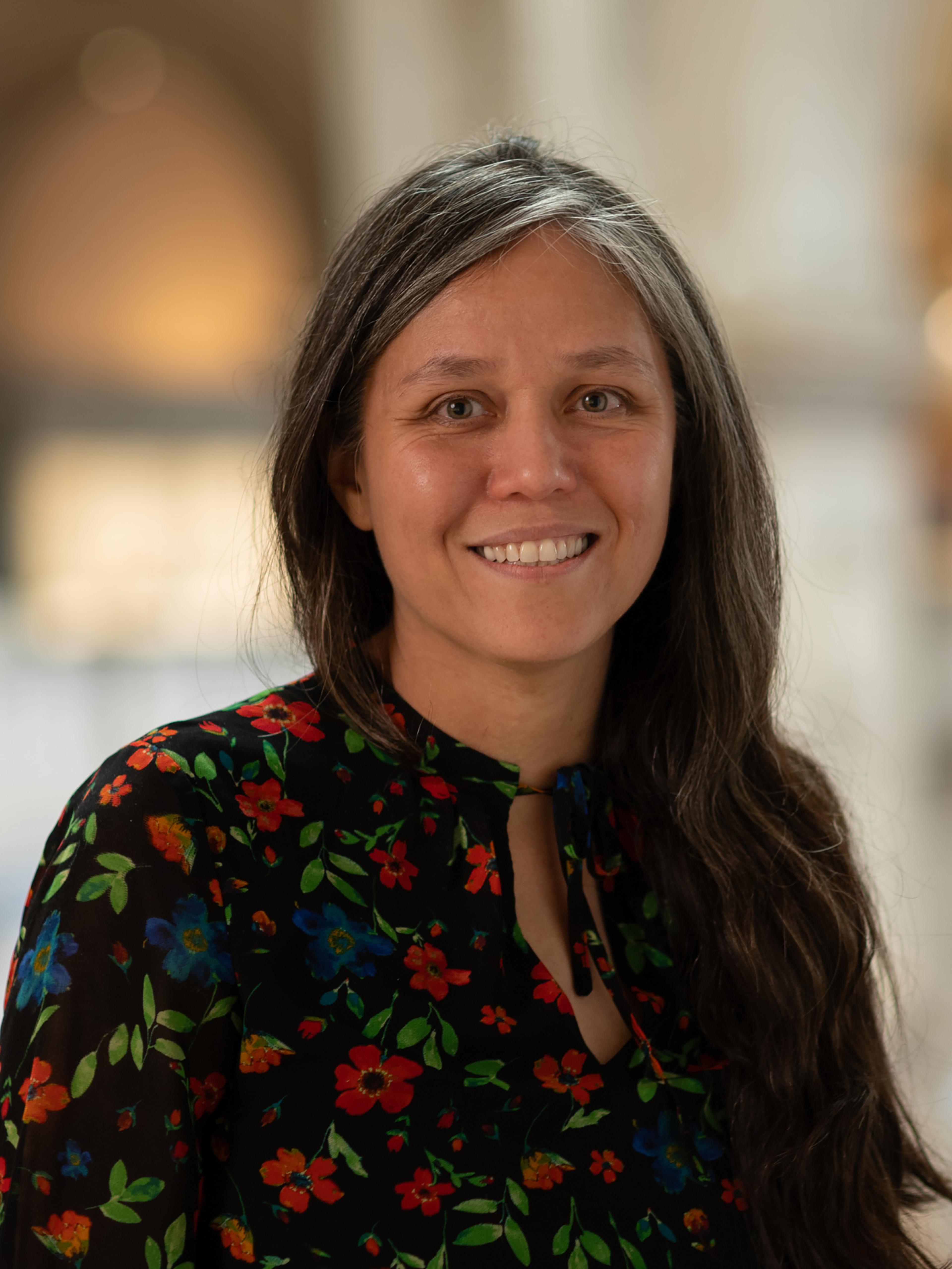 Headshot of Liz Zanis. She is smiling and looking directly at the photographer with long dark hair and a long sleeve dark top with a red flower pattern. 
