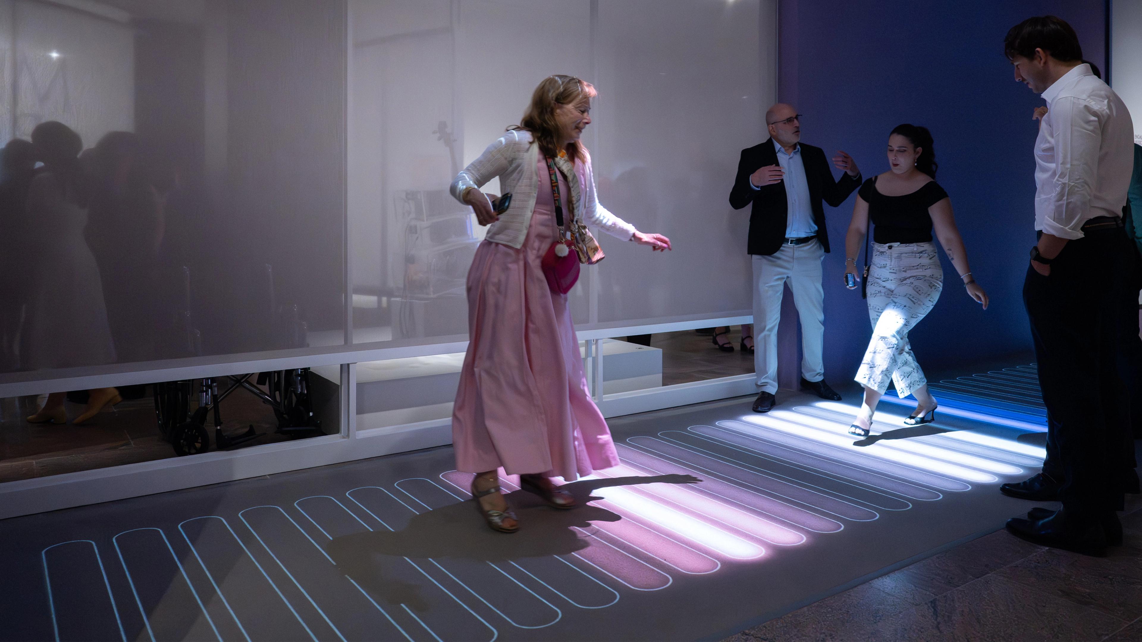 A woman in a pink dress interacts playfully on a glowing, colored floor panel, surrounded by people in a dimly lit room