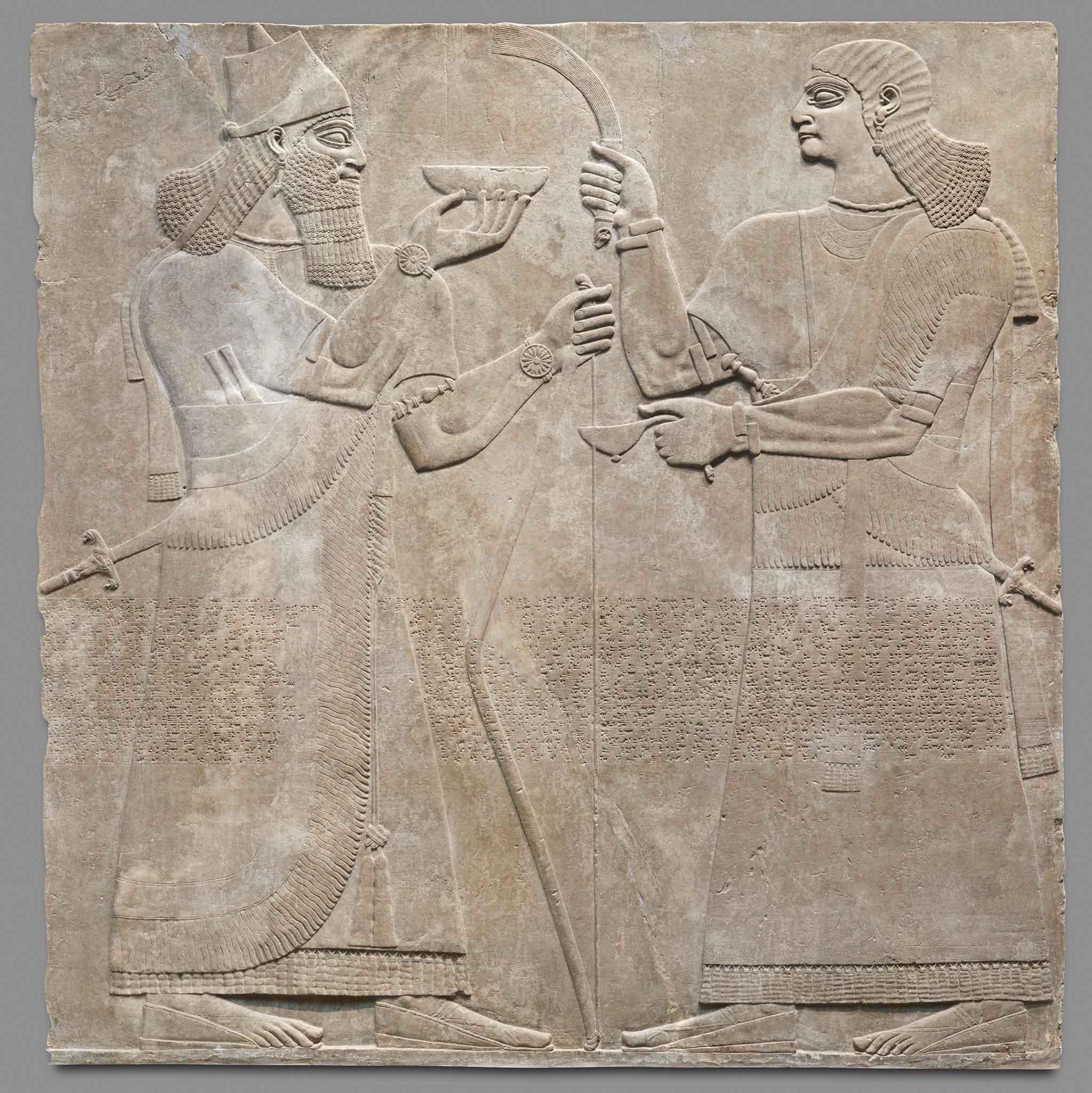 Assyrian relief panel showing a king holding a bowl, and his attendant at right. Both are richly dressed and carry swords.