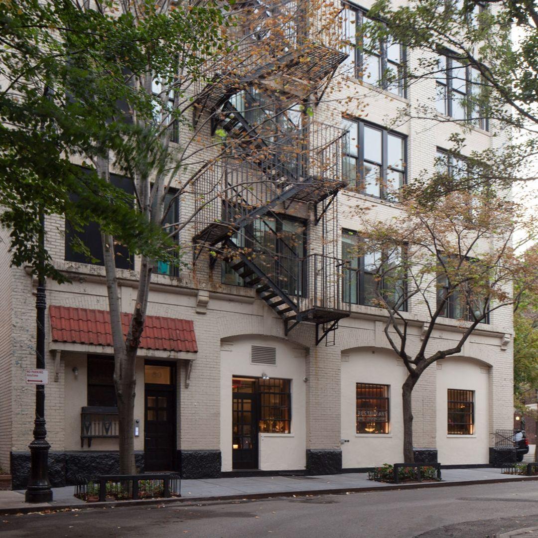 Contemporary photograph of a multi-story building with a brick facade painted beige with black fire escapes and a narrow tree-lined sidewalk.
