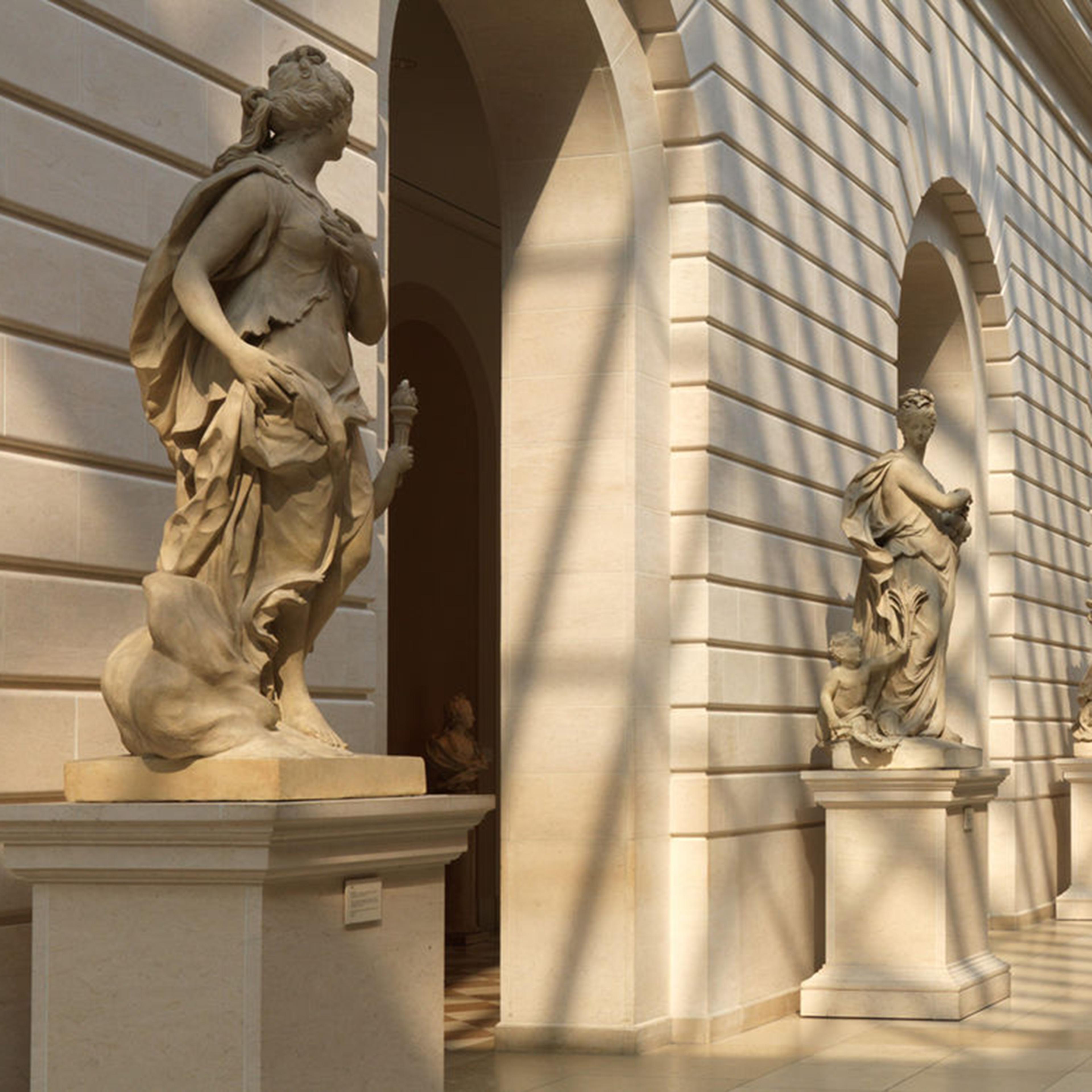 A sunlit gallery featuring classical sculptures against elegant stone walls, with large windows allowing natural light to flow in.