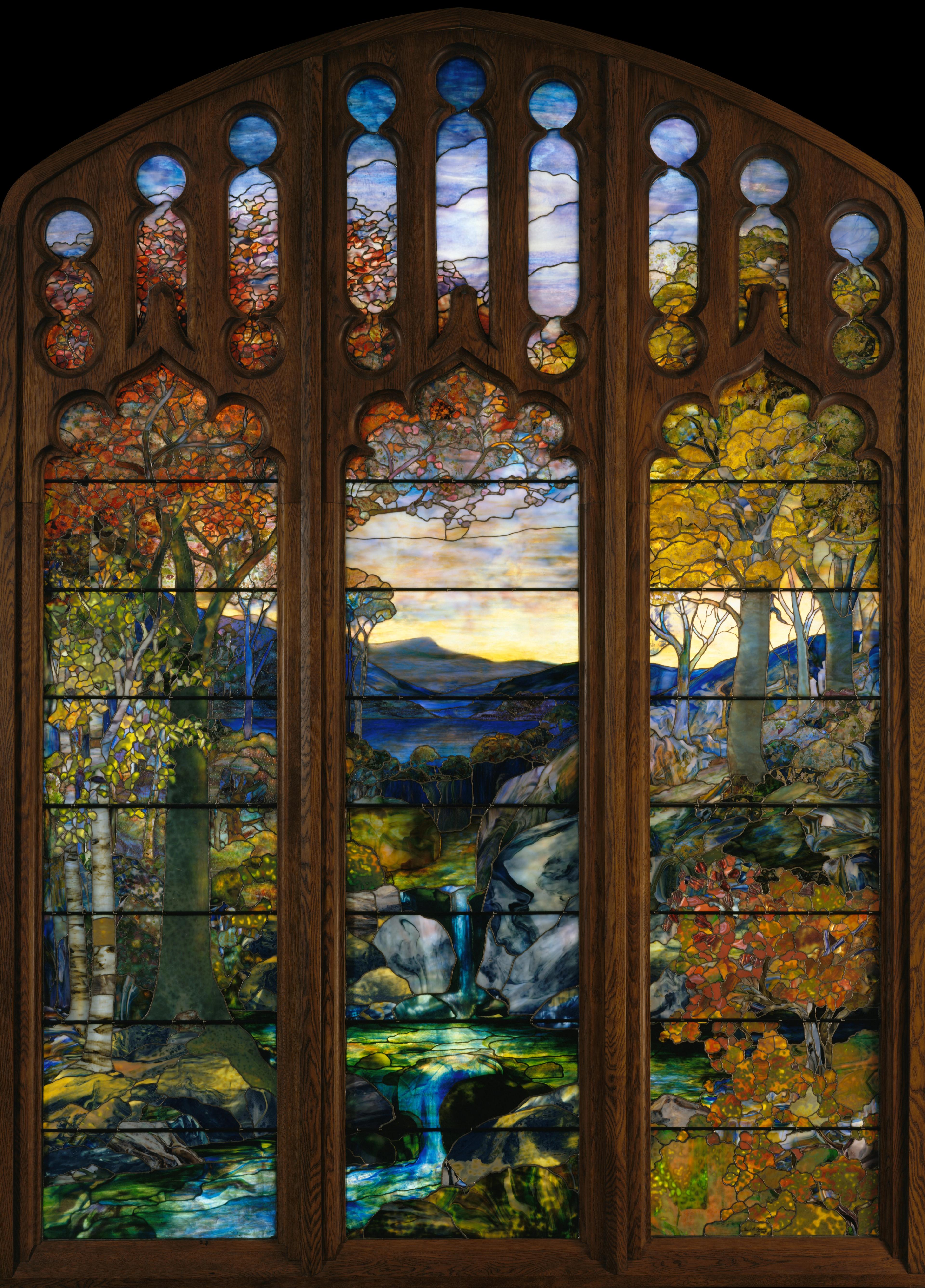 A tall, colorful stained-glass window depicting a forested landscape with flowing water, autumnal trees, and distant mountains.