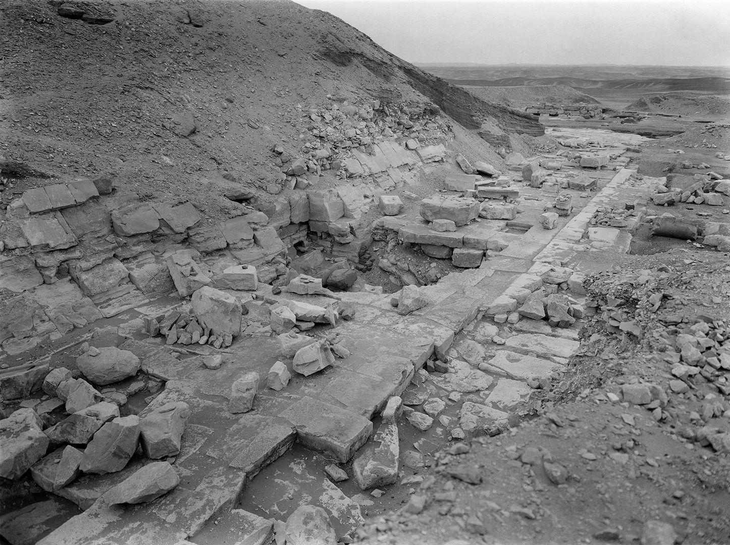 Black and white view of the side of a partially destroyed pyramid to the left with part of a floor surface of slabs of stone along its edge