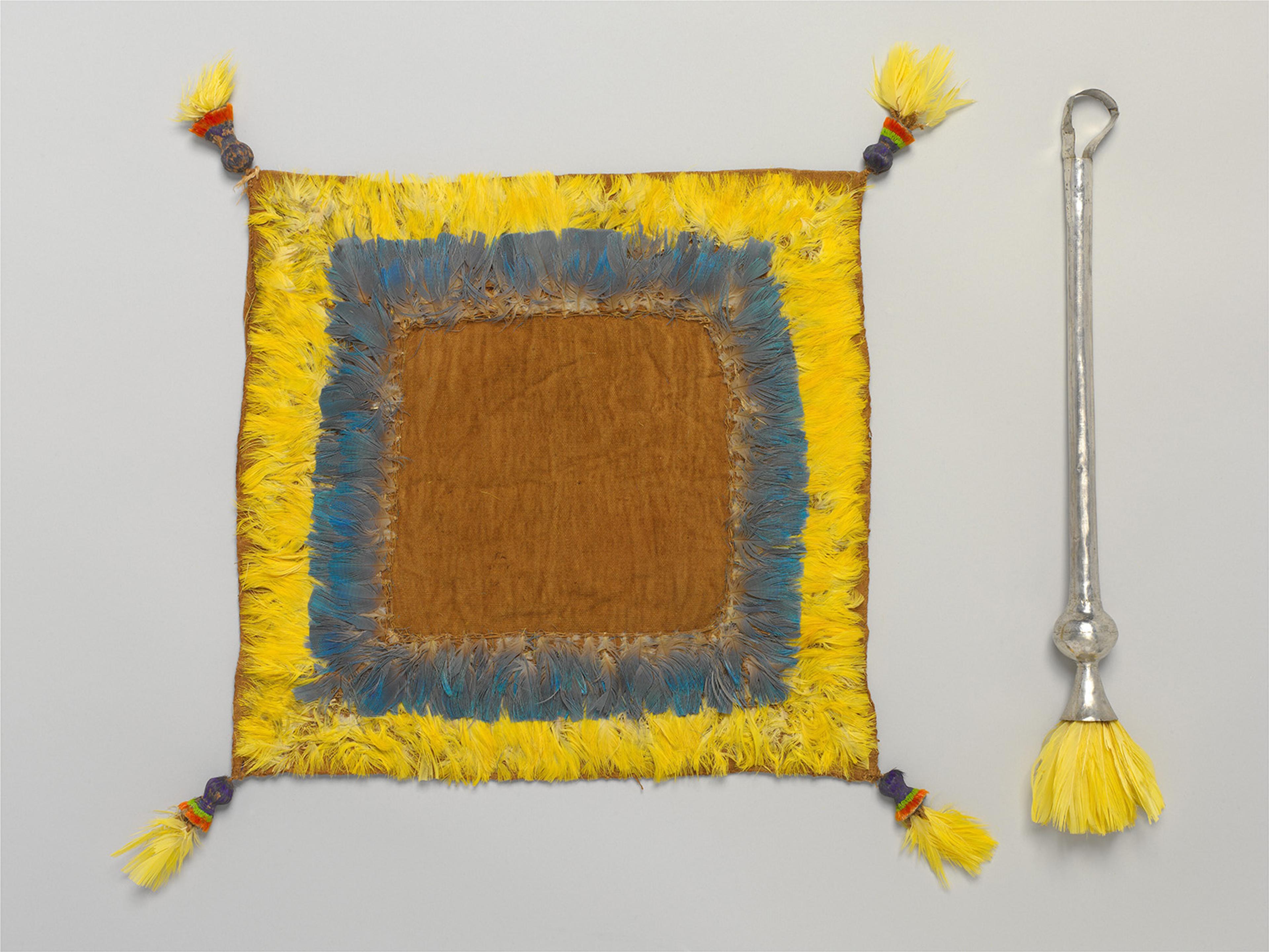 Handwoven square textile with yellow, blue, and brown concentric bands, corner tassels, and a silver needle-like tool with yellow bristles below.
