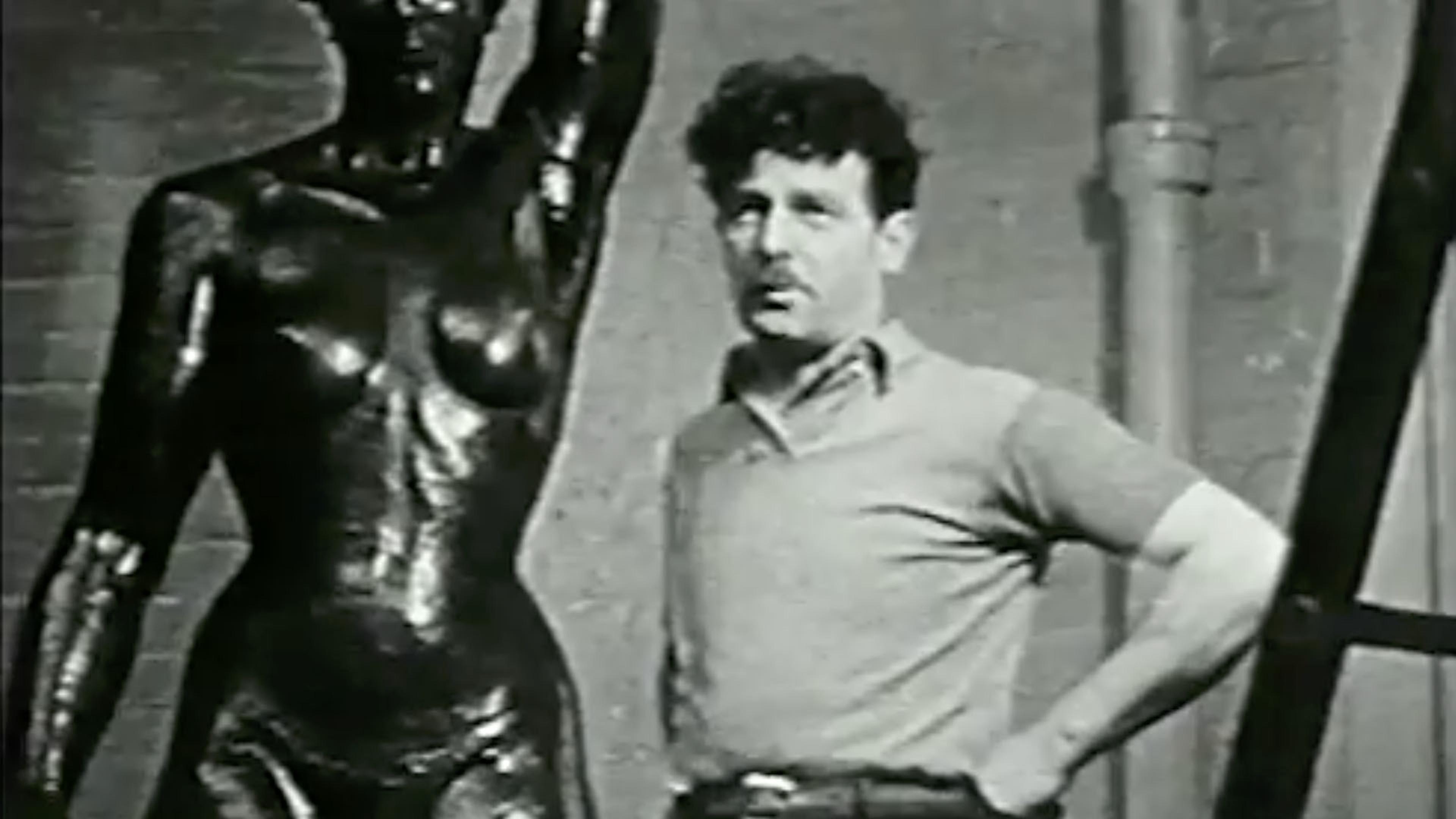 A black and white video still of a man standing next to a sculpture of a female nude
