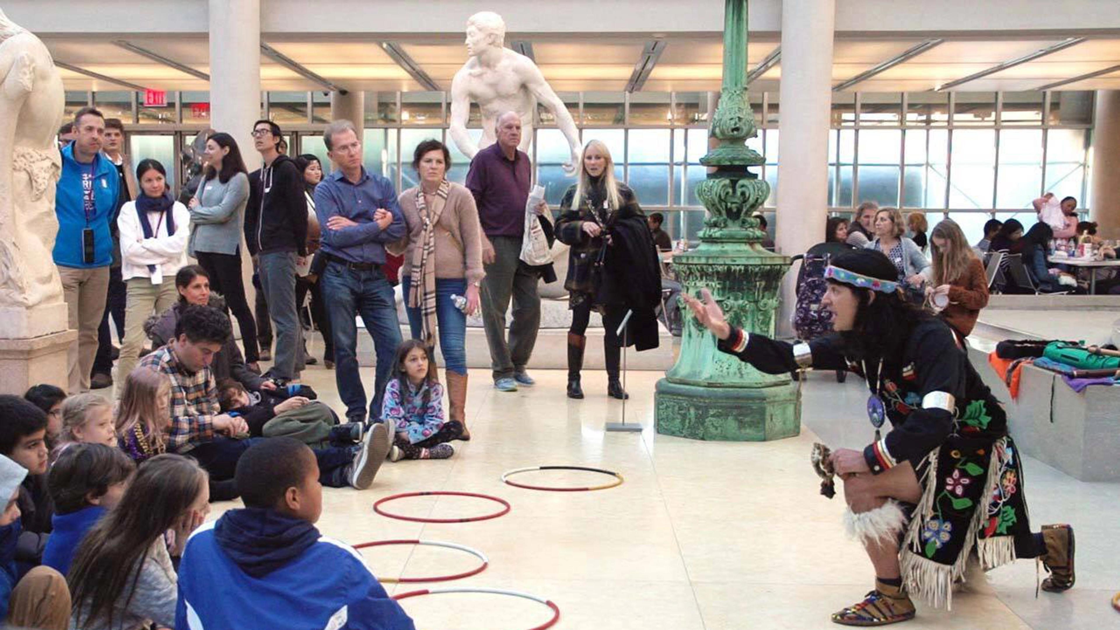 Artist Ty Defoe kneels in front of a group of children and parents in The Met's American Wing.