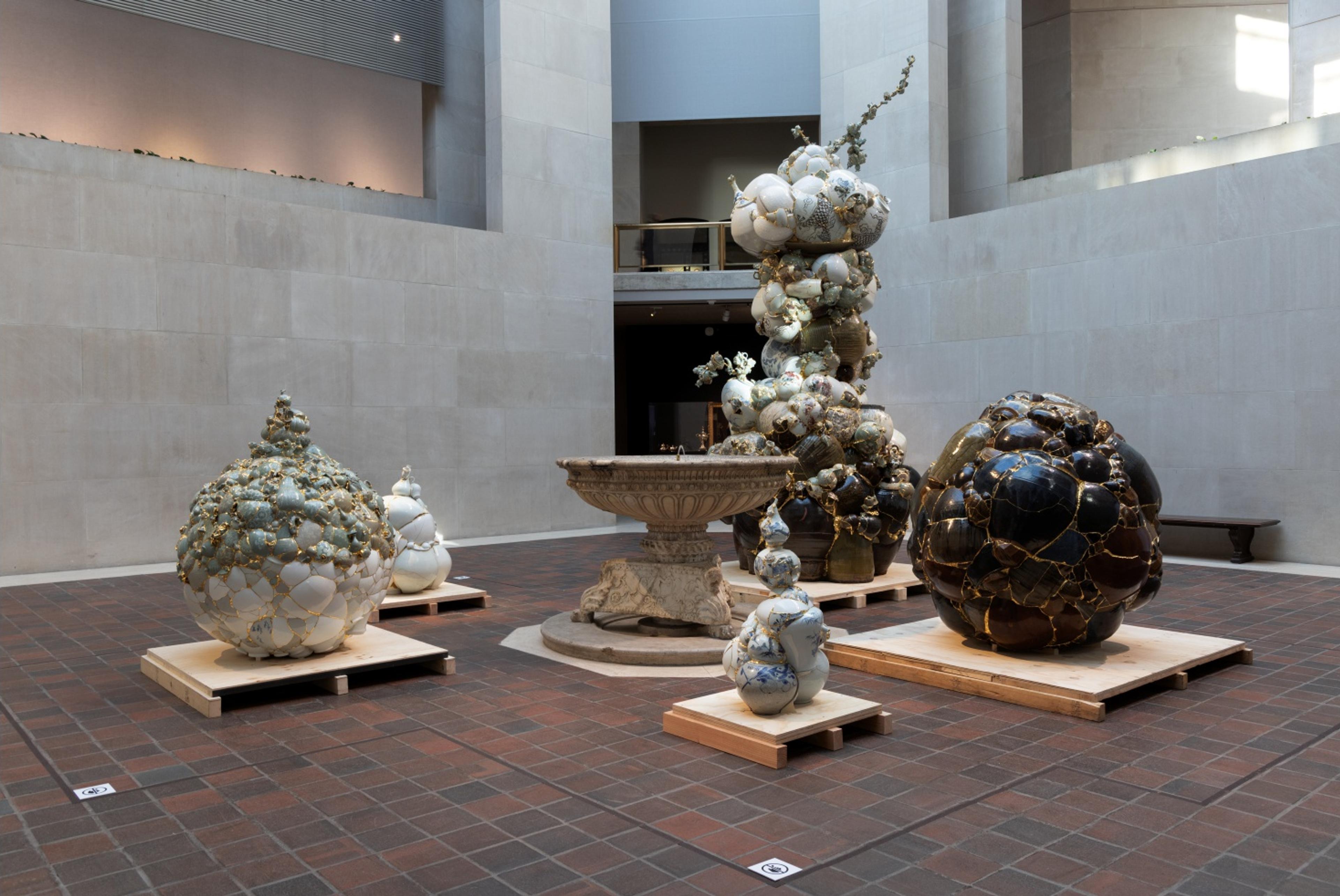 Exhibit of five large ceramic sculptures, resembling clustered spheres with gold accents, surrounds a stone fountain in a spacious, light-filled gallery.