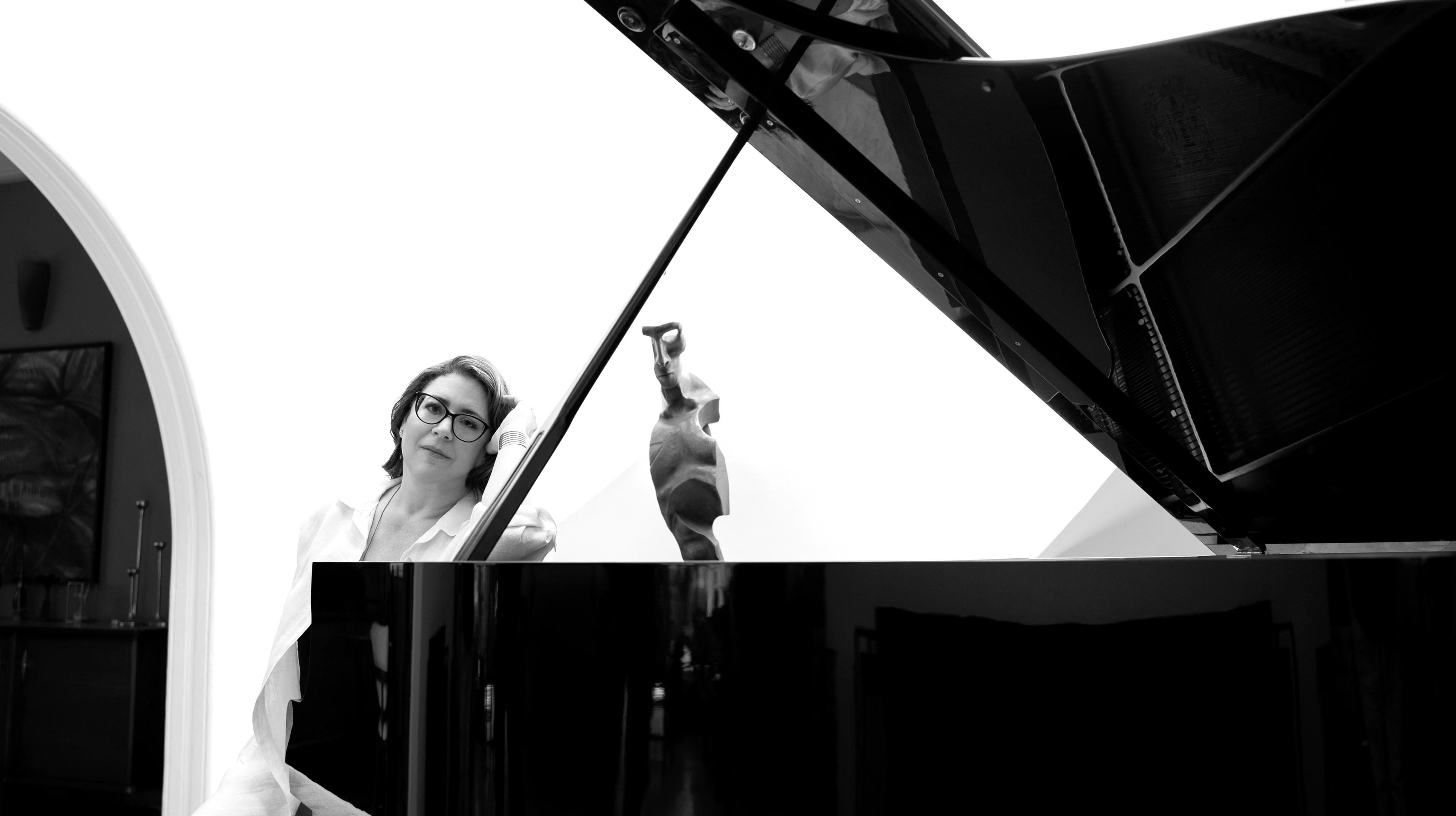 A woman poses behind a grand piano, leaning her head against her hand.