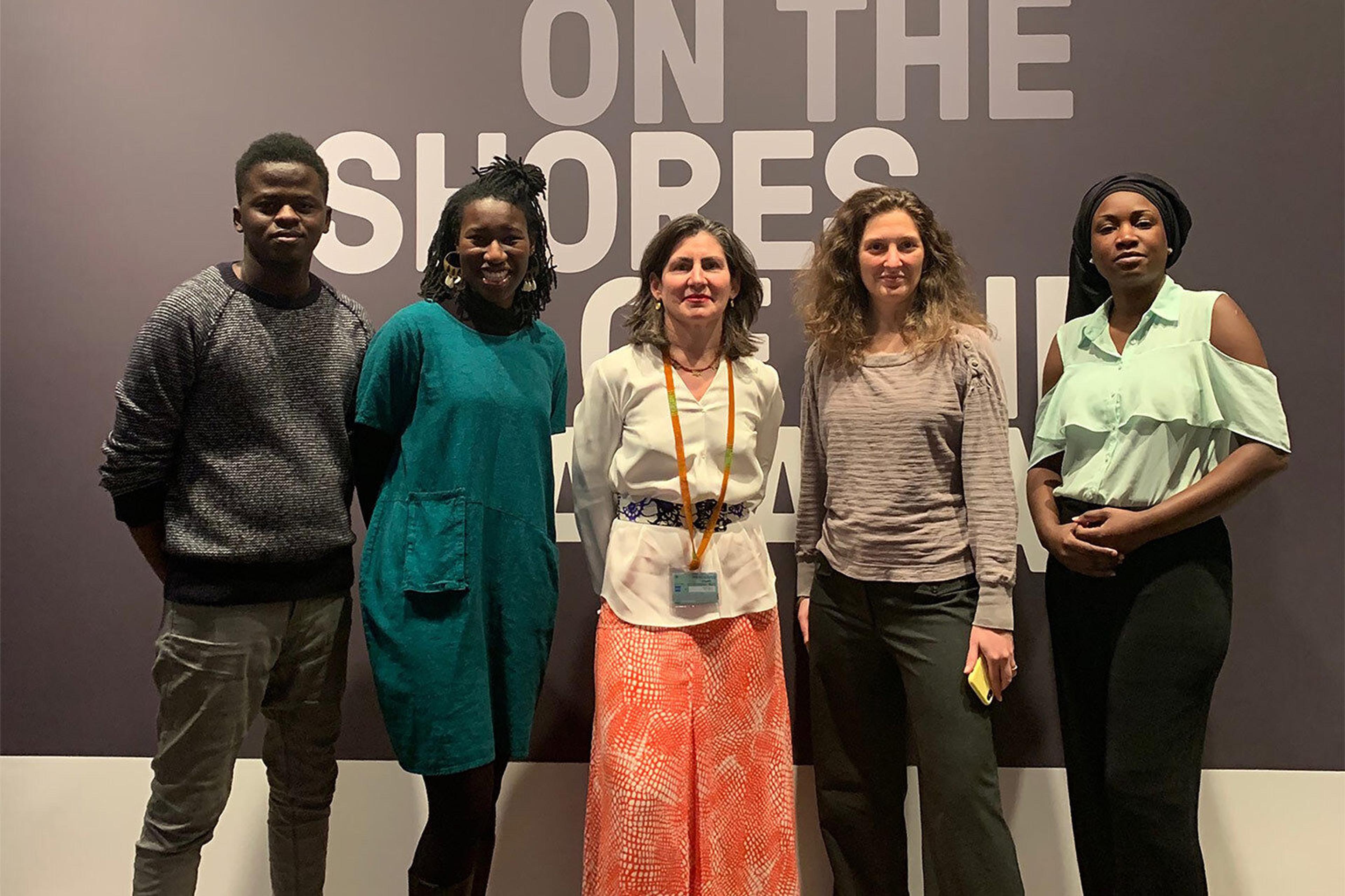Five people smile and stand in front of an ivory wall with white text that reads: "On the Shores"