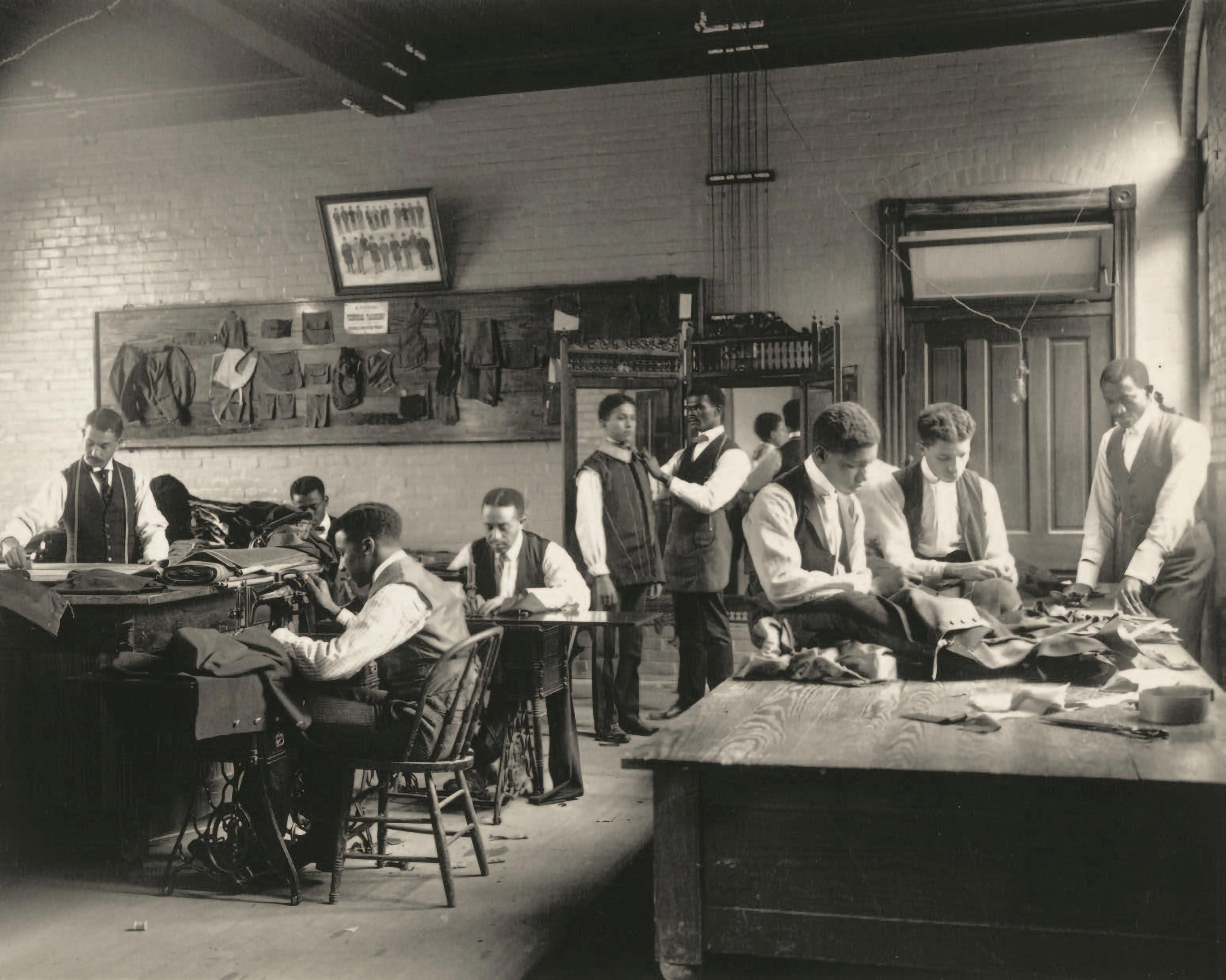 Black-and-white photograph of a tailoring studio. Young men sit sewing around a table at left, while another group stands, observing fabrics laid out on a table at right