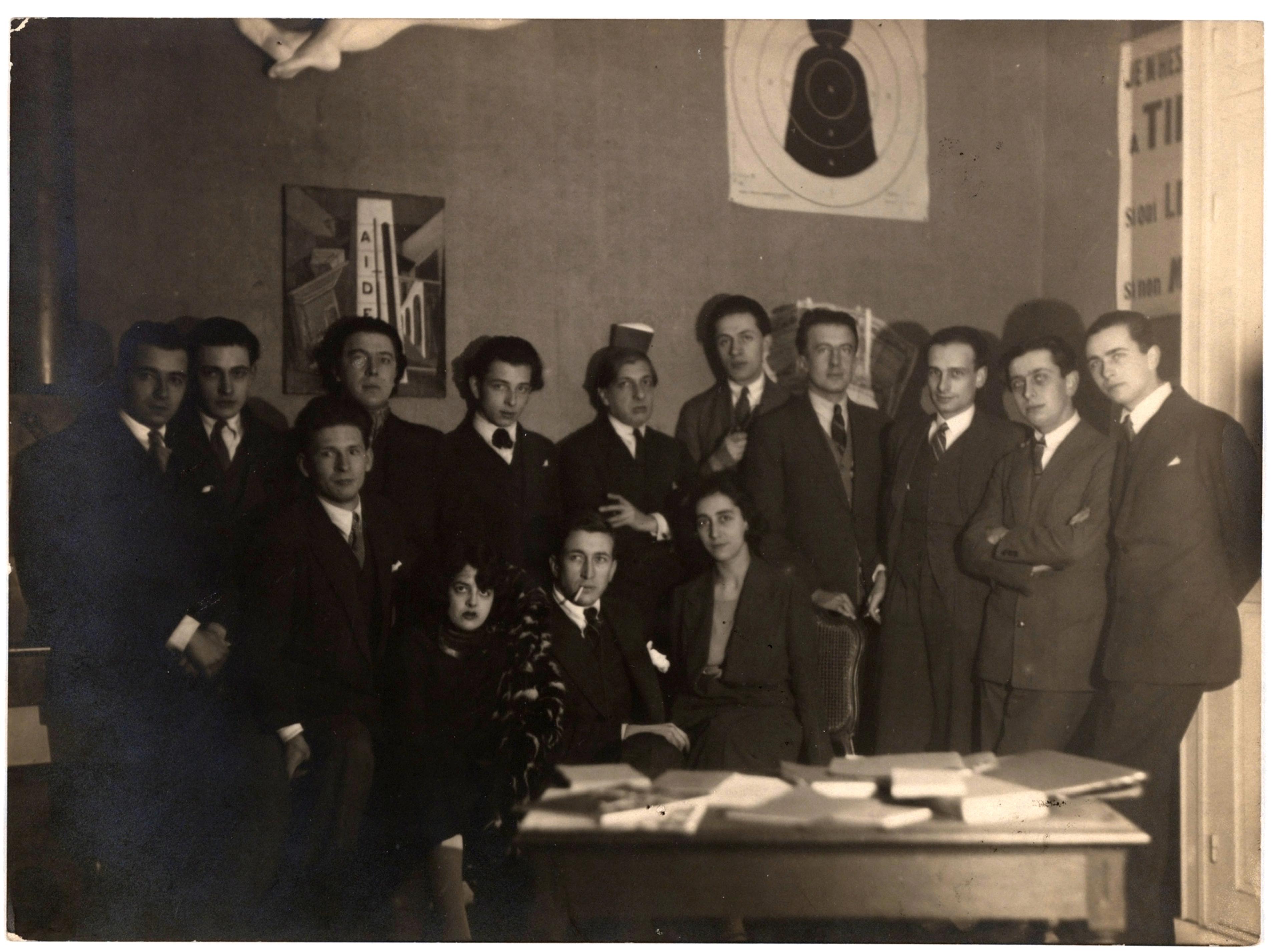 Black-and-white portrait of a group of people wearing dark suits in front of a desk covered in books and papers.