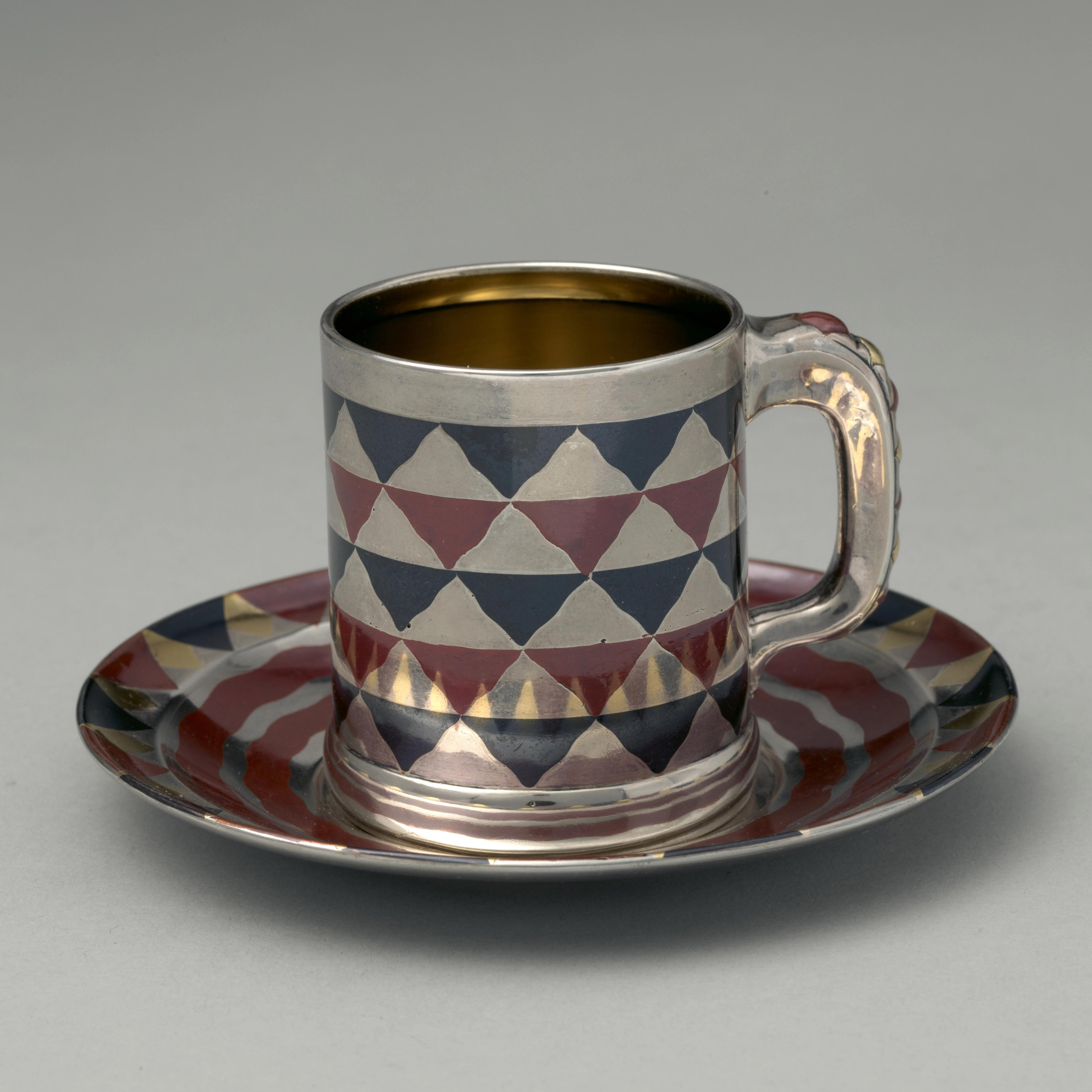 A silver, blue, and maroon cylindrical cup with a handle rests on a matching saucer. The cup and saucer are reflective and adorned with an inlay design of a checkerboard pattern of wavy triangle shapes.