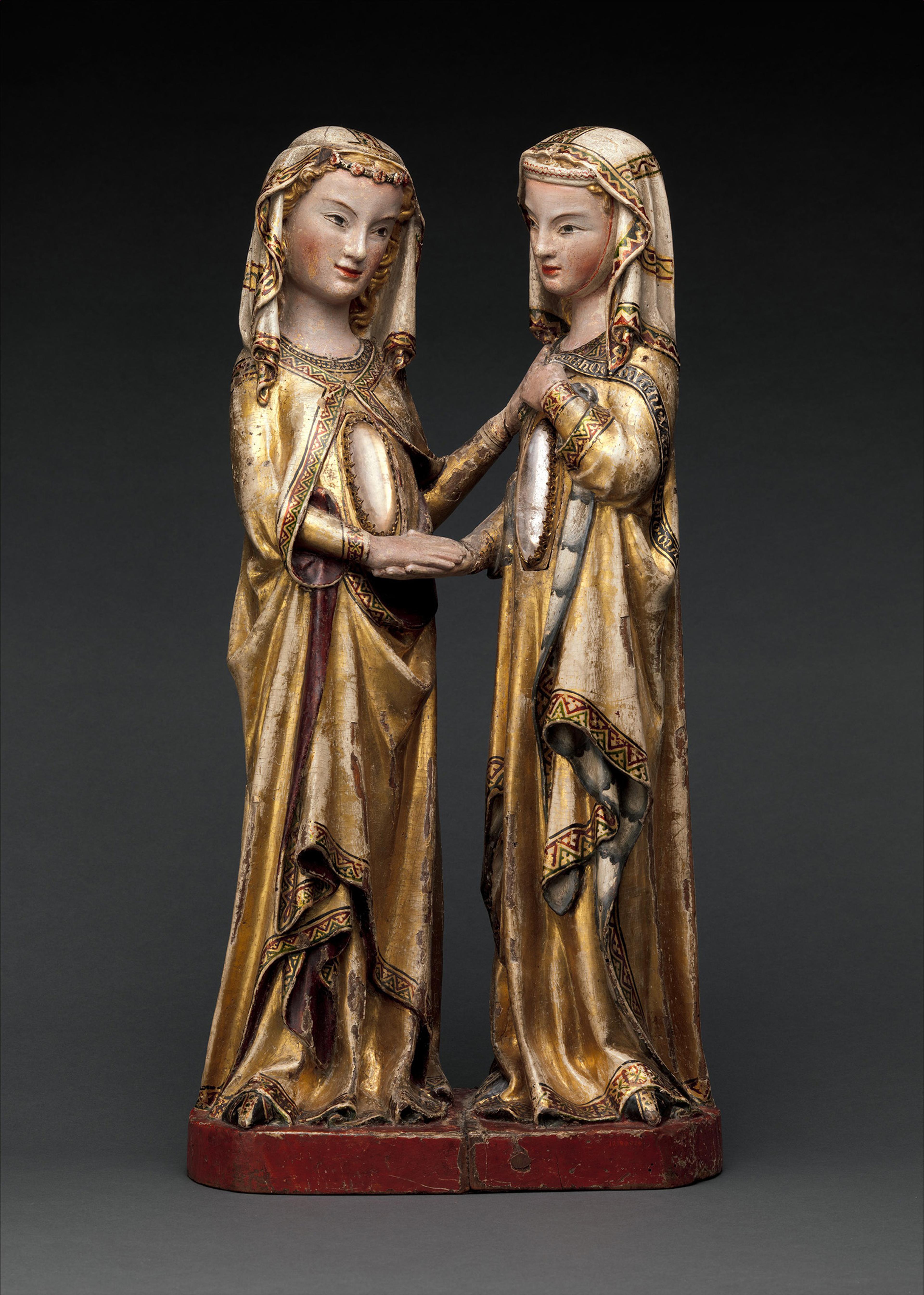 Two intricately carved figures in medieval robes and veils stand facing each other, hands clasped in a gesture of connection, against a dark background.