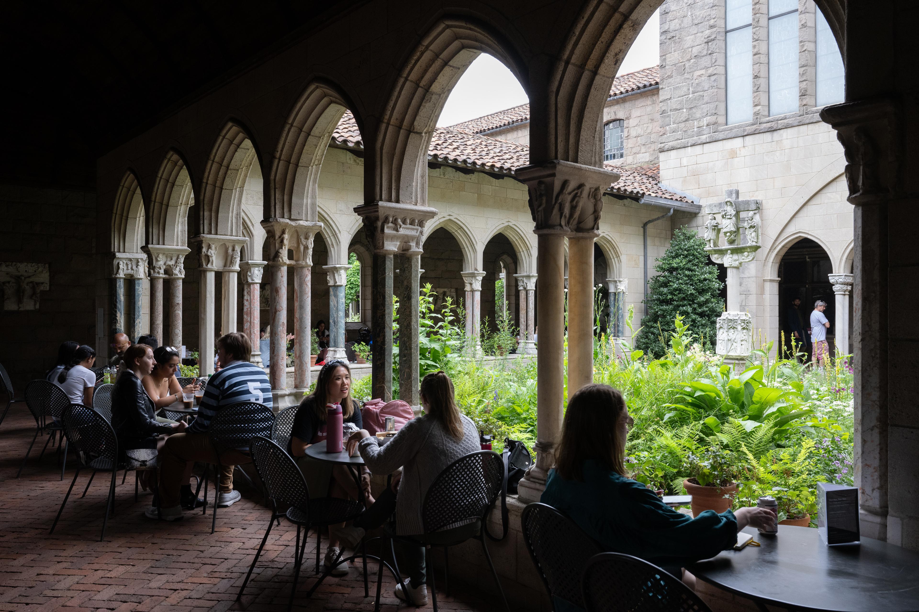 Patrons sit at tables and chairs in a stone arcade overlooking a garden.