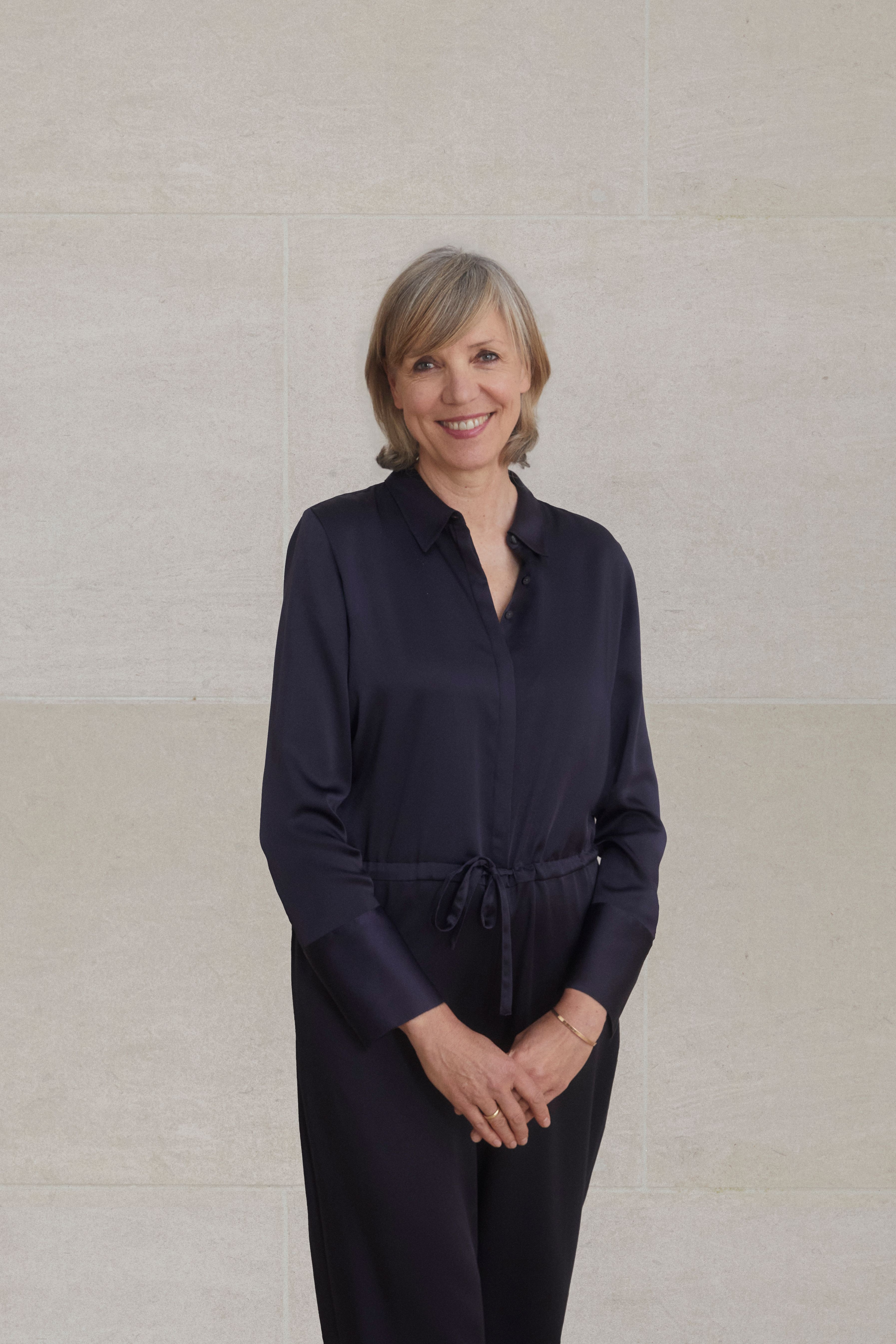 A color photograph of Inka Drogemuller standing in front of a stone wall in a black top and pants.