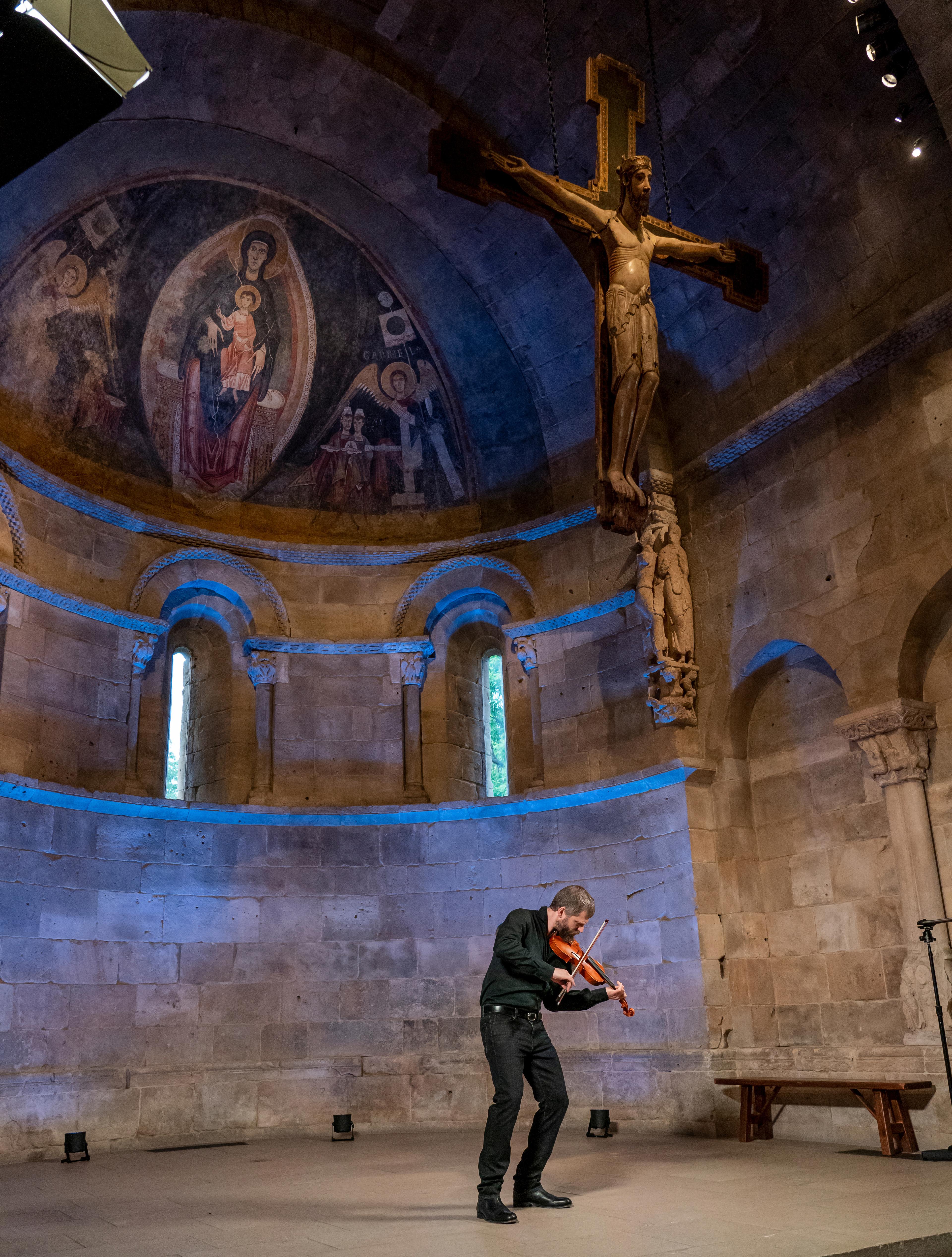 Johnny Gandelsman in performance at The Met Cloisters. He is dressed in black, violin tucked under his chin. The ceiling above features frescoes and a large cross.