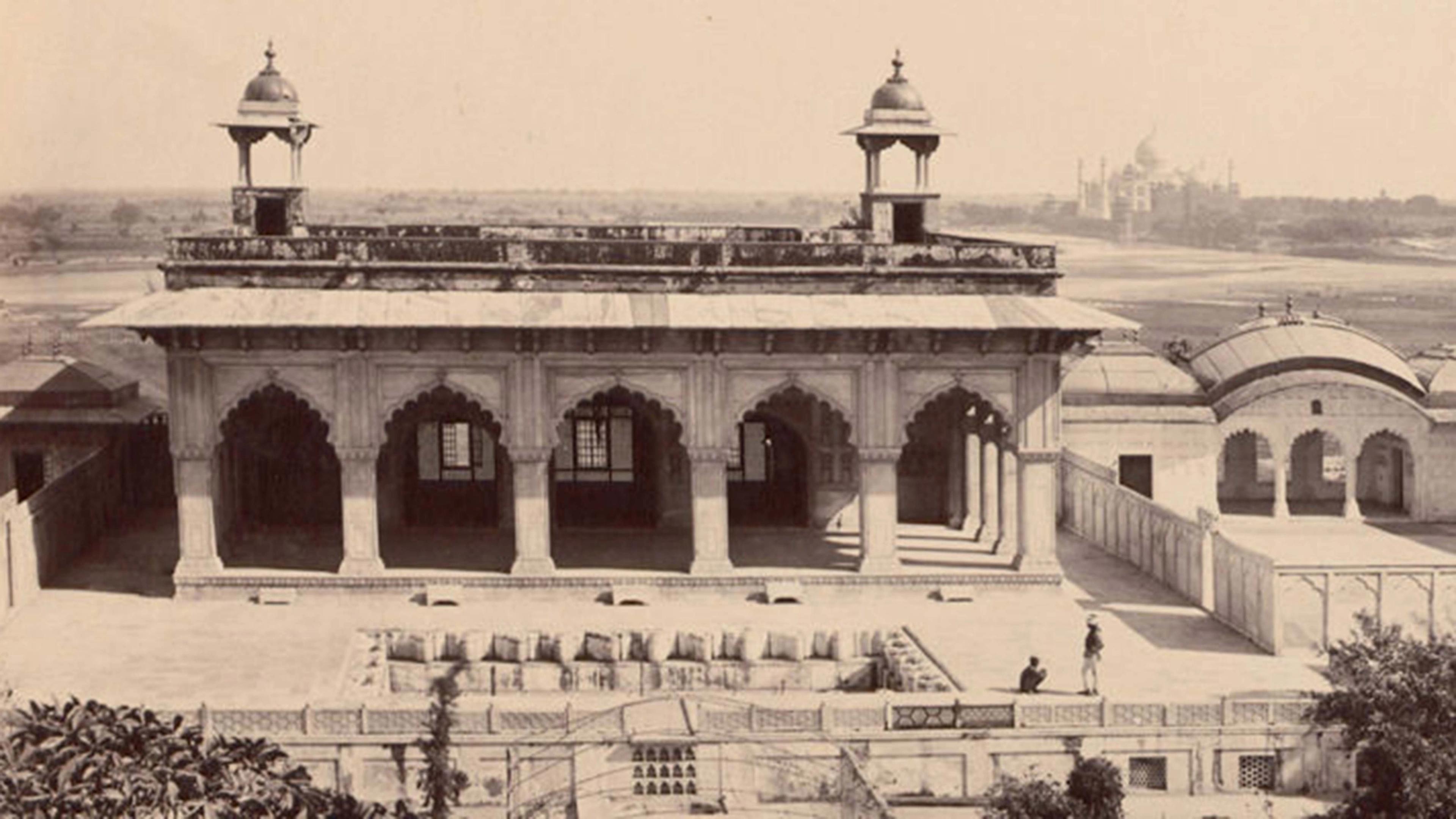 A historic building with arched columns and two domed pavilions, with the Taj Mahal visible in the background across an open landscape