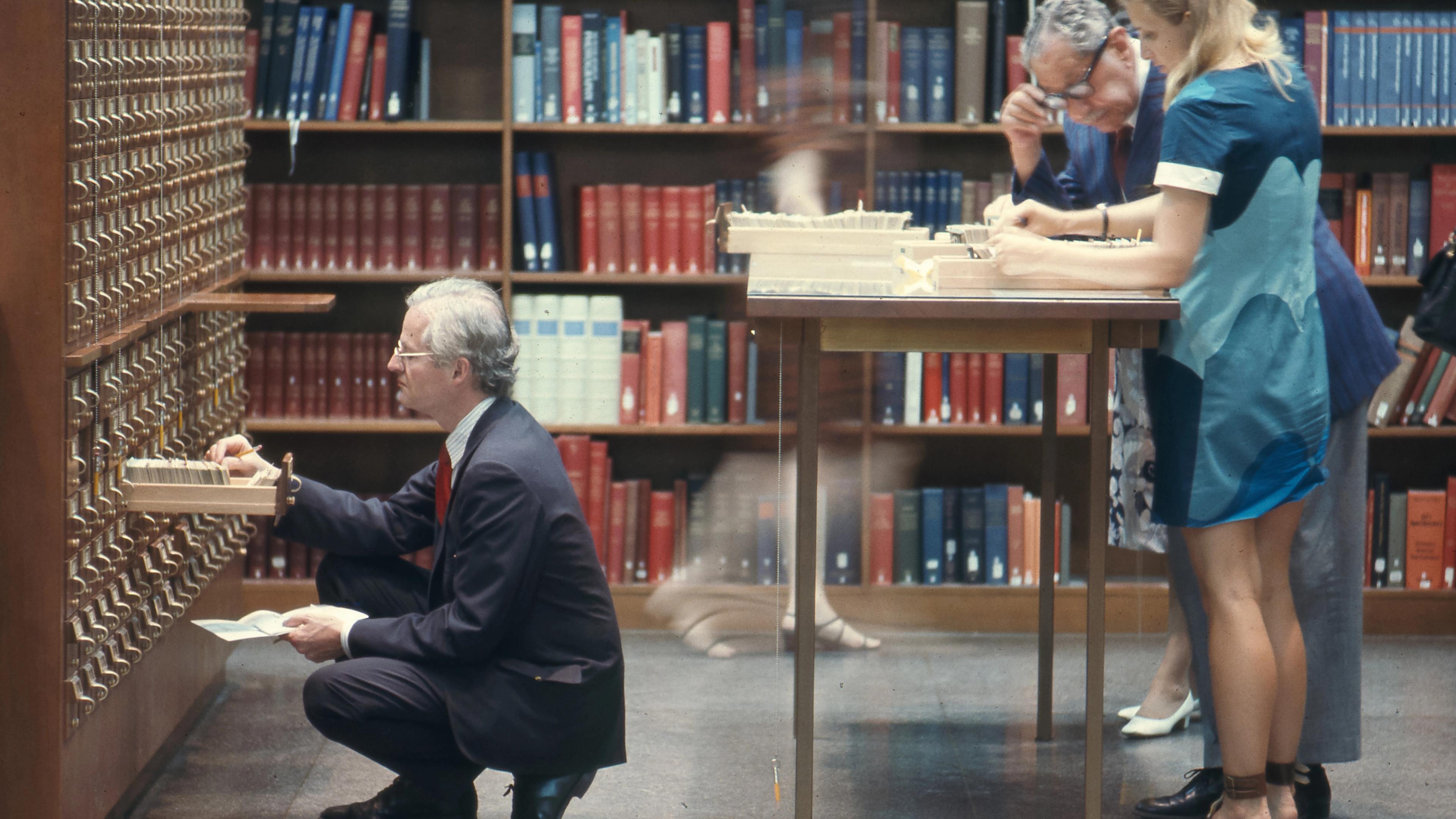 Dated photograph of people using a library card catalog and reference files, with bookshelves in the background.