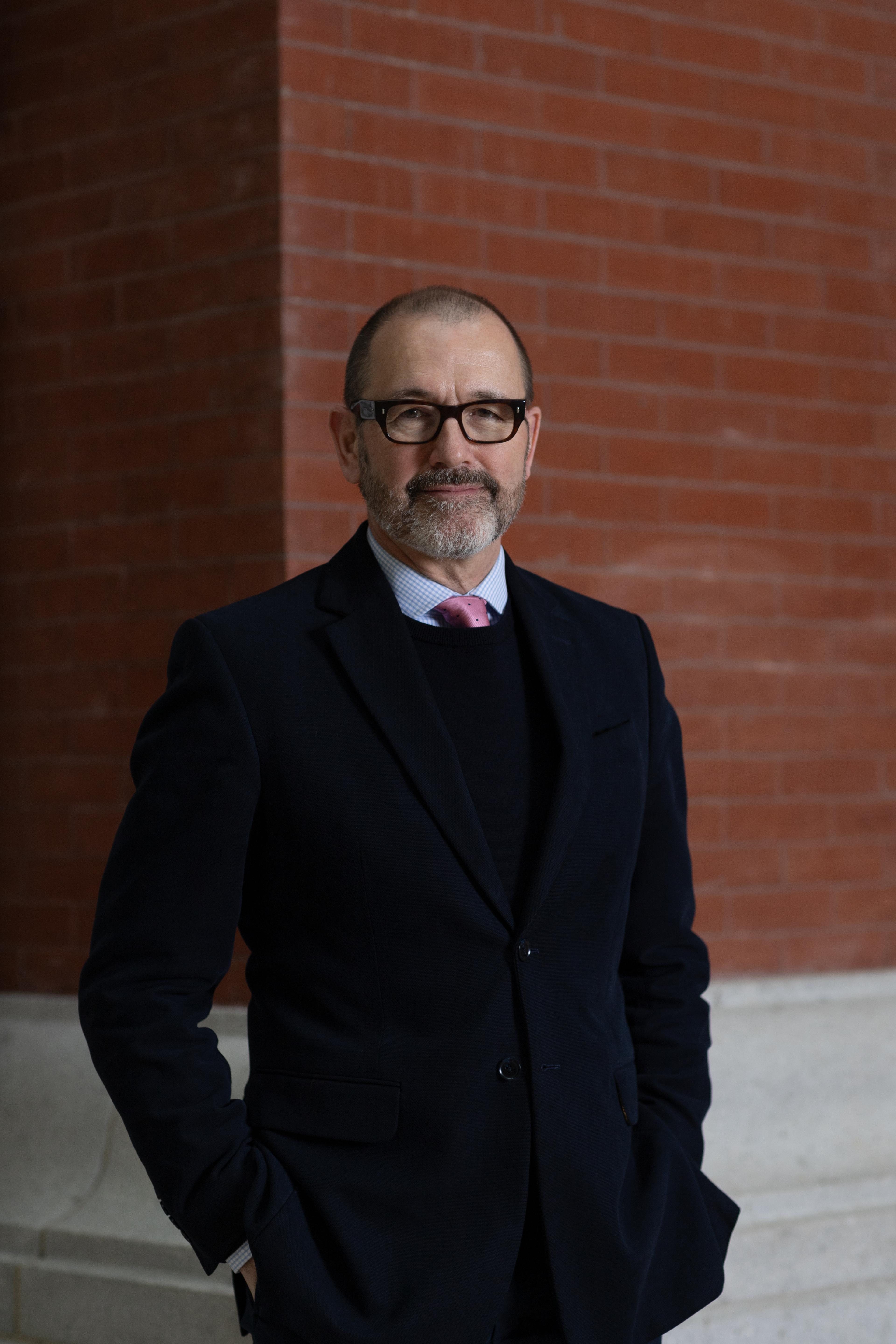 Head of Lucian Simmons in a dark suit, dark sweater, and pink tie standing in front if a red-brick wall.  