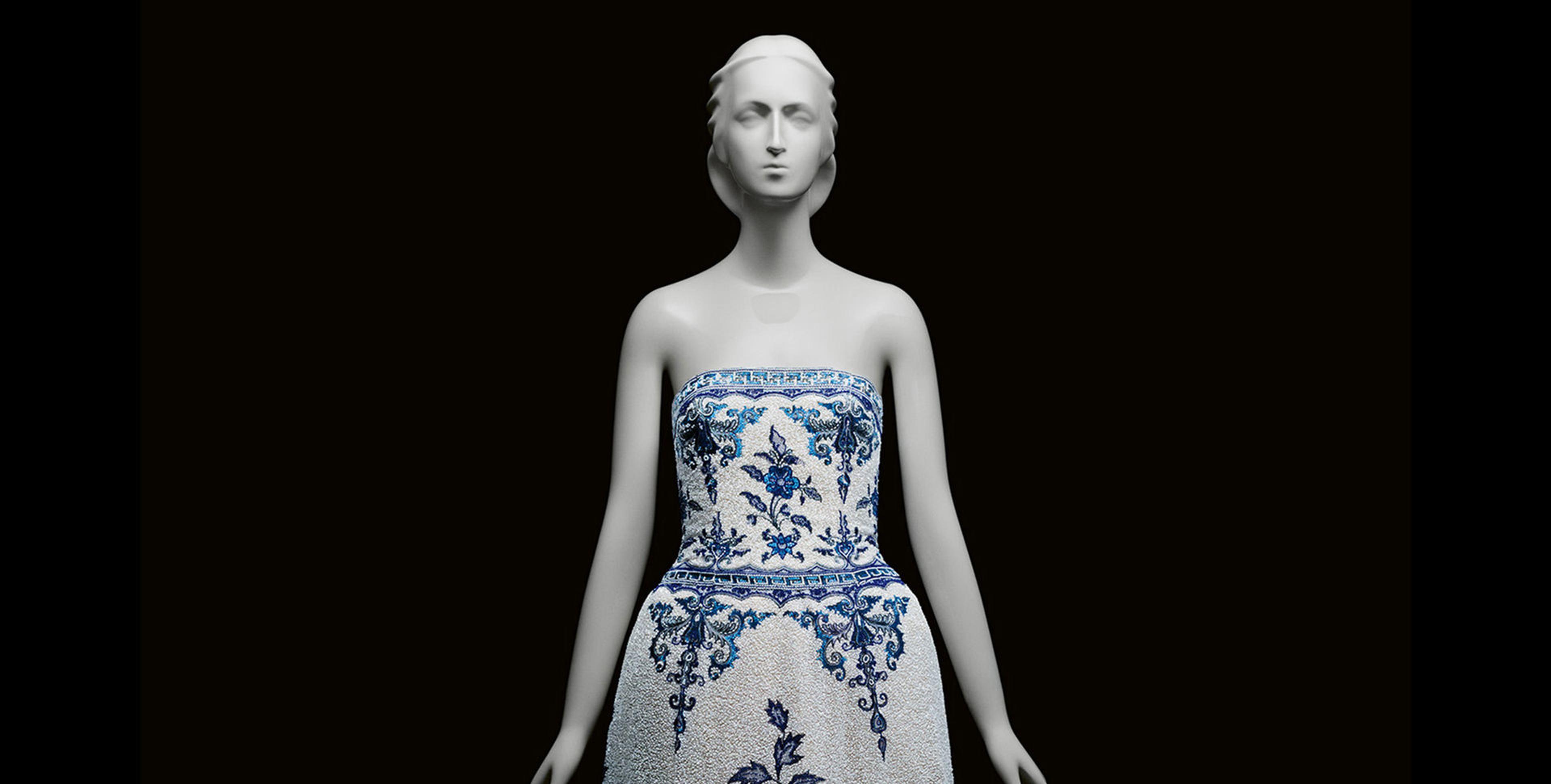 The back of a navy blue dress with beautiful white patterning on the back and lower part of the dress and around the wrists. The dress is against a grey background and fitted onto a mannequin with a head and hands.