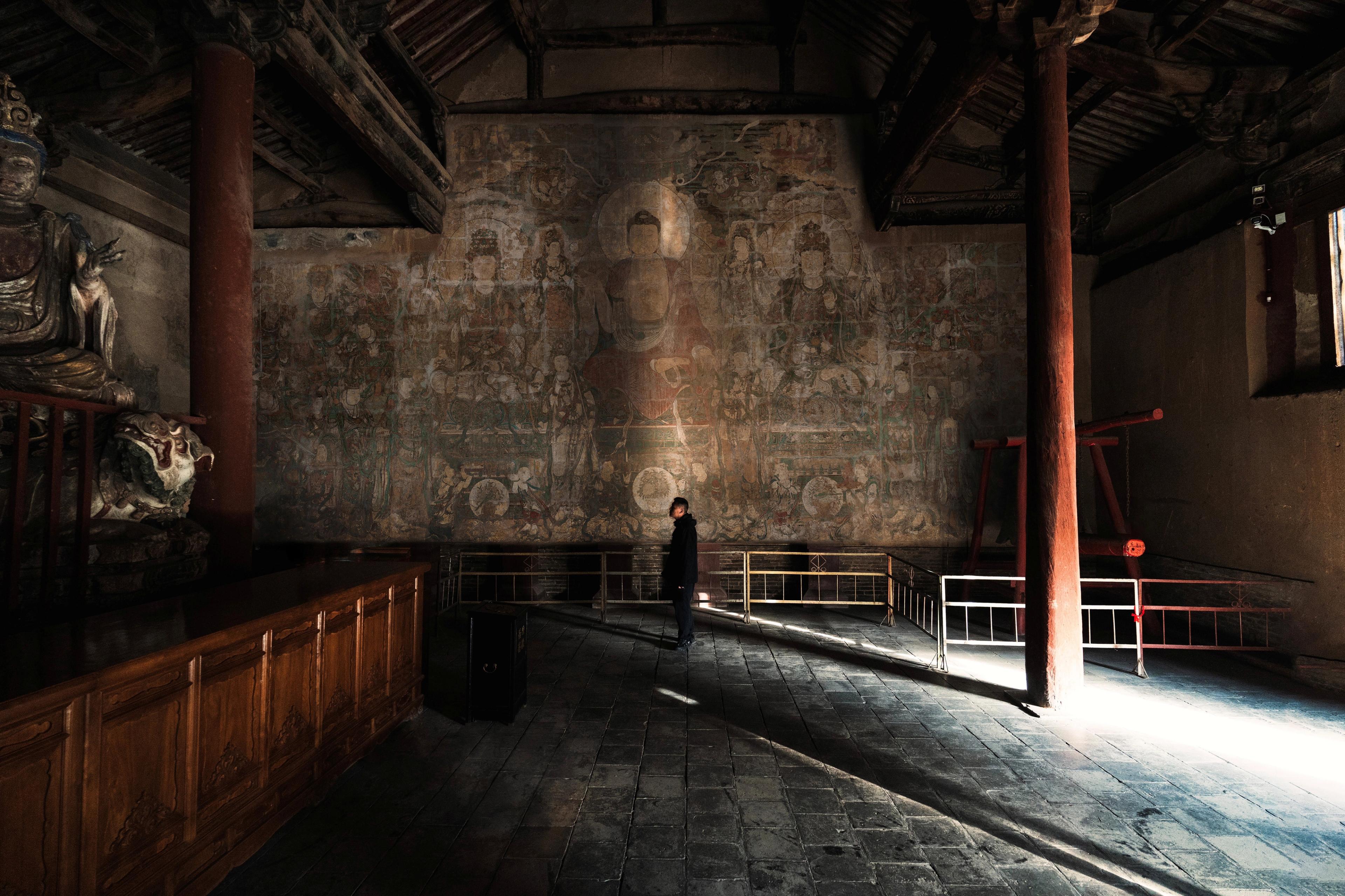 Light streams in as a single bright beam to a dimly lit temple space with a Buddha painting on the far wall.