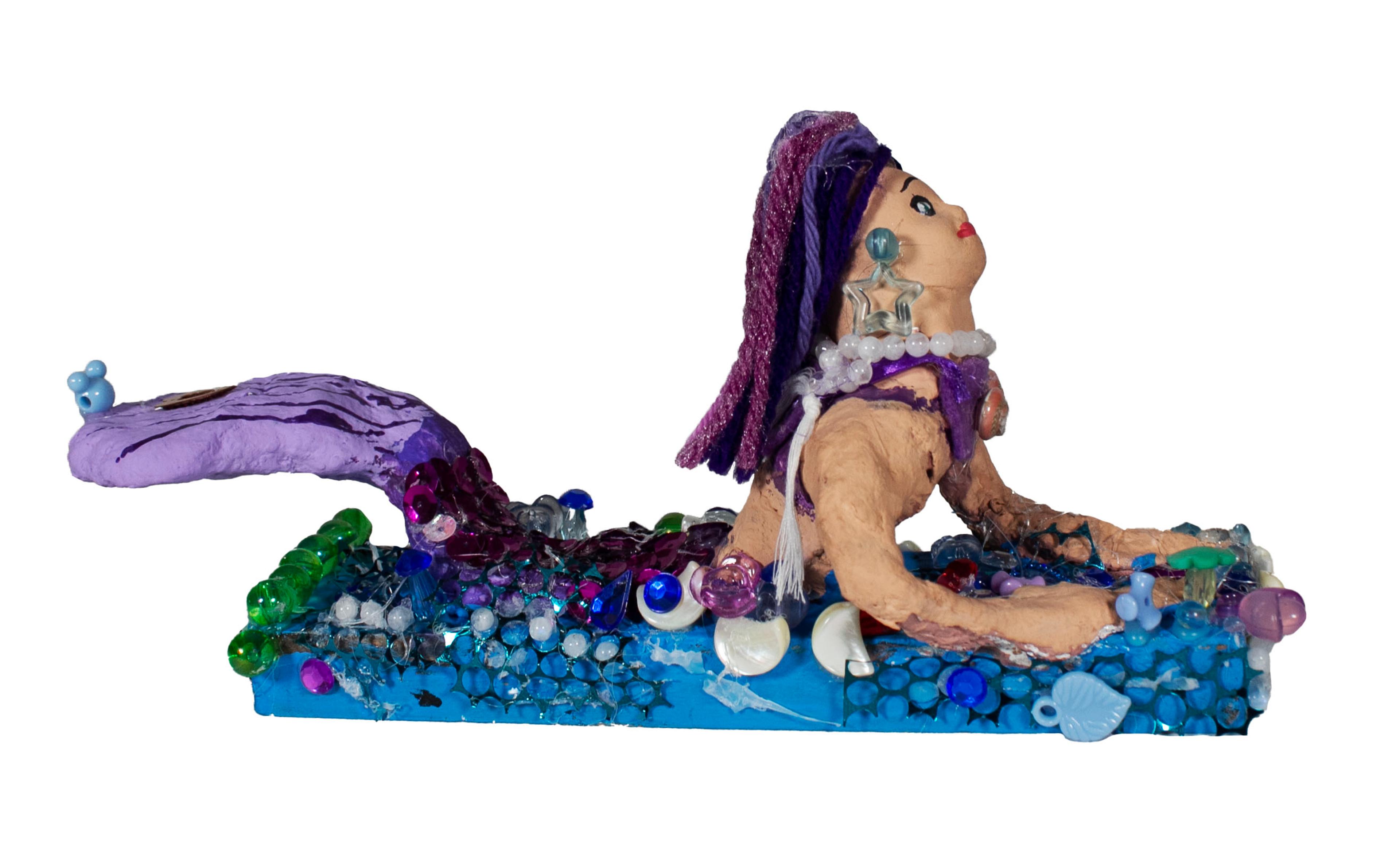 Mixed-media sculpture of a mermaid emerging from the frothy rectangular surface of a blue body of water. The mermaid faces right, looking upward, with her arms resting on the surface of the water. The mermaid's purple tail lifts up from the water to the left. She wears a transparent, star-shaped earring and has purple braided hair. She wears a purple necklace underneath a white pearl necklace. The mermaid is surrounded by various shiny and colorful baubles that suggest seashells from the ocean.