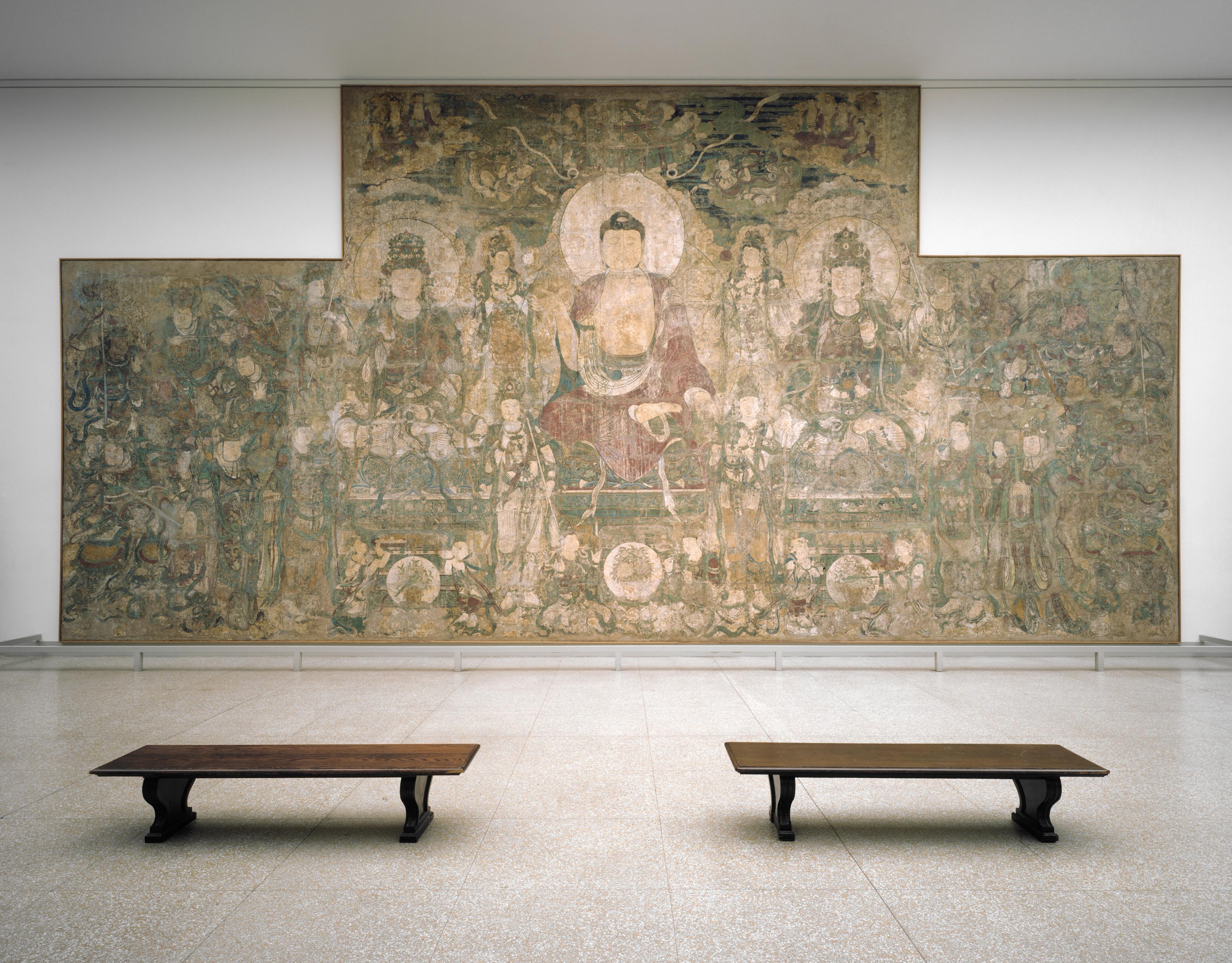 Large painting of Bhaishajyaguru, the Buddha of Medicine, sitting on a lotus throne surrounded by an assembly of deities, installed in a gallery with two benches in the foreground.
