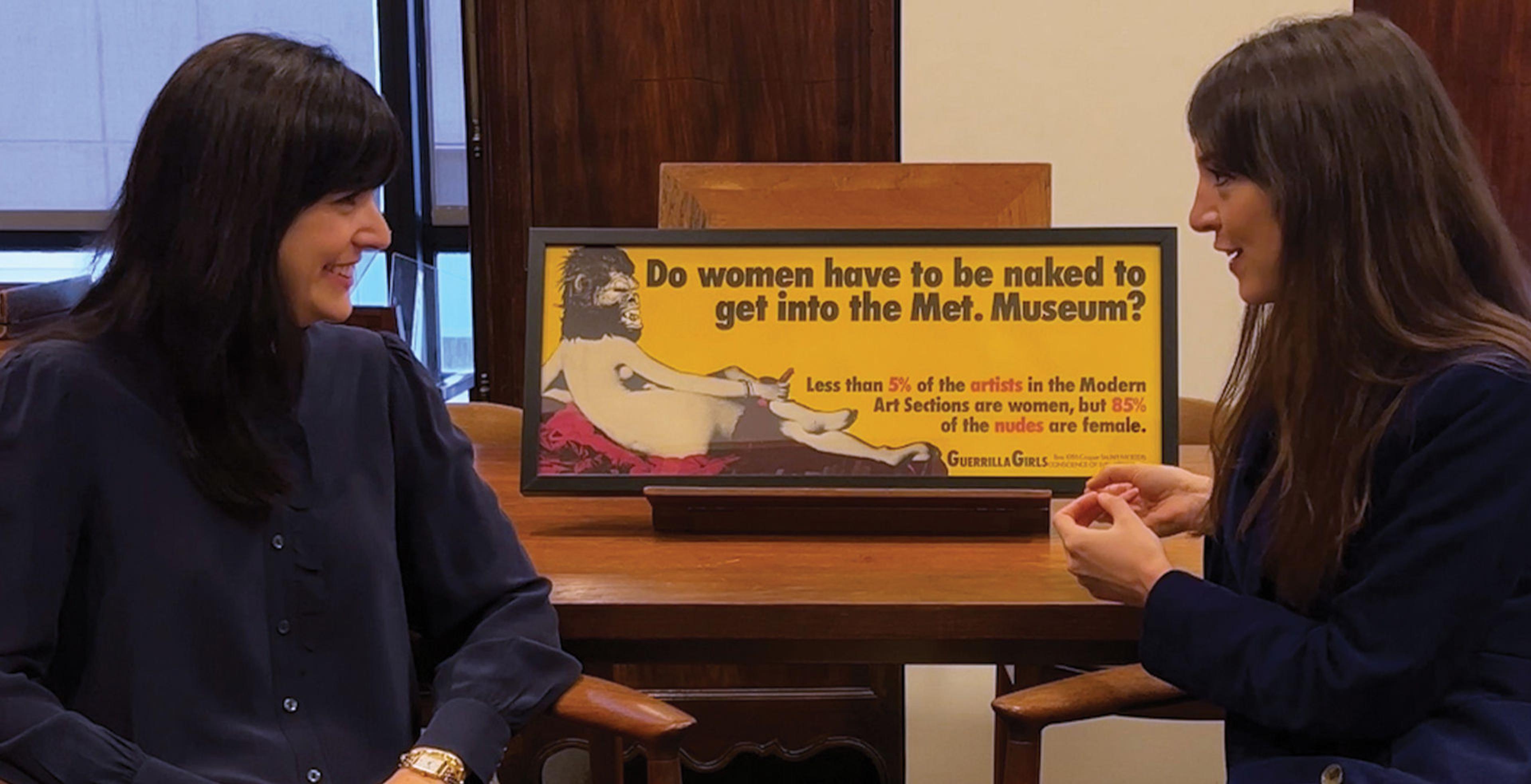 Allison Rudnick and Katy Hessel speak in front of of the print artwork "Do Women Have To Be Naked To Get Into the Met Museum?" by the Guerrilla Girls.