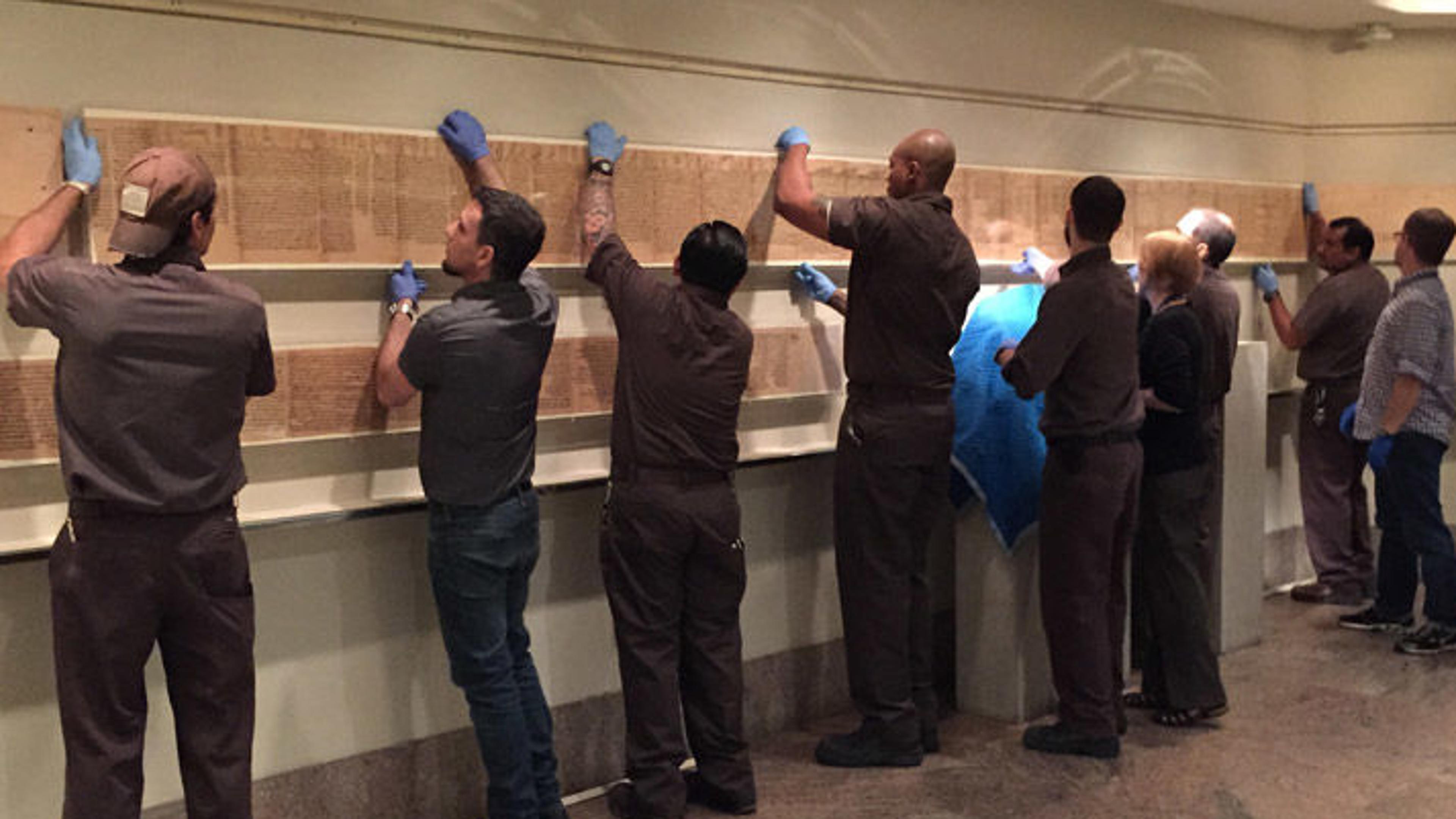 A row of workers holding up a long scroll along a gallery wall.