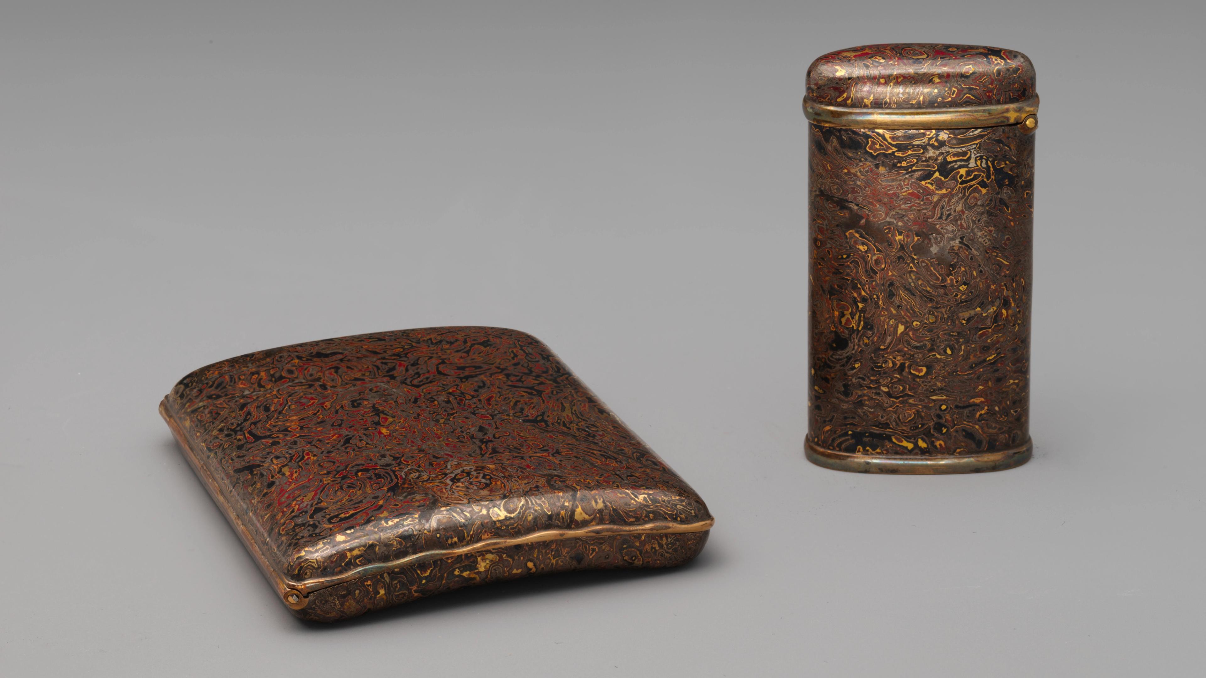 A flat cigarette case and a tall match case in a matching wood grain pattern rendered in deep red, black, brown, and gold metals. 