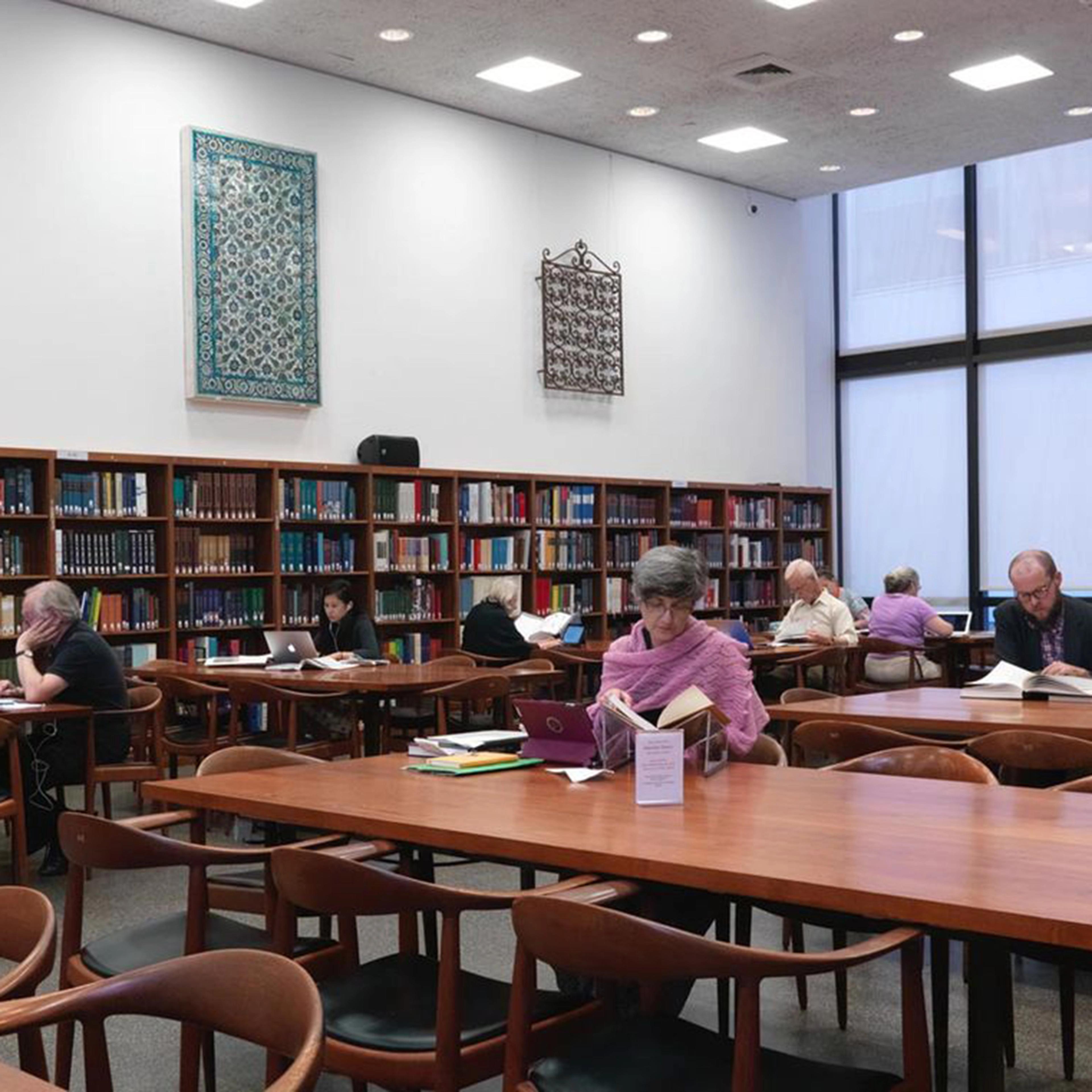 Individuals seated at large wooden desks reading with a wall of bookshelves and windows in the background.