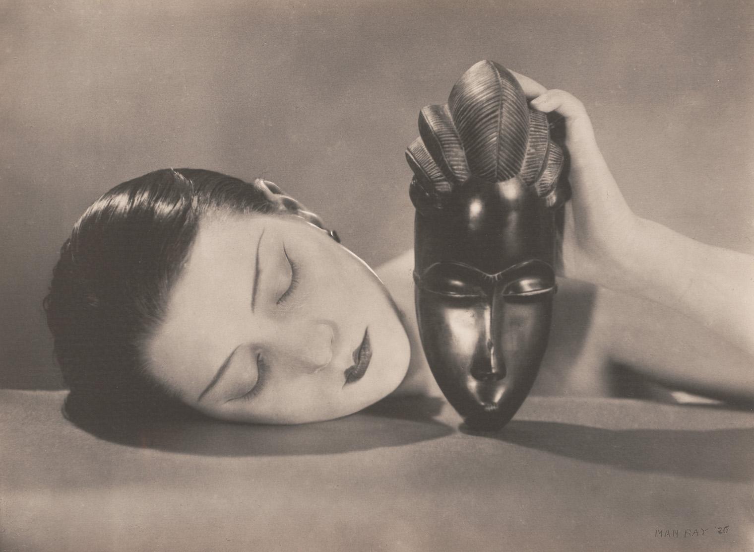 A black and white photo shows a woman resting her head on a surface with closed eyes, holding an African-style mask beside her face. The mood is serene and contemplative.