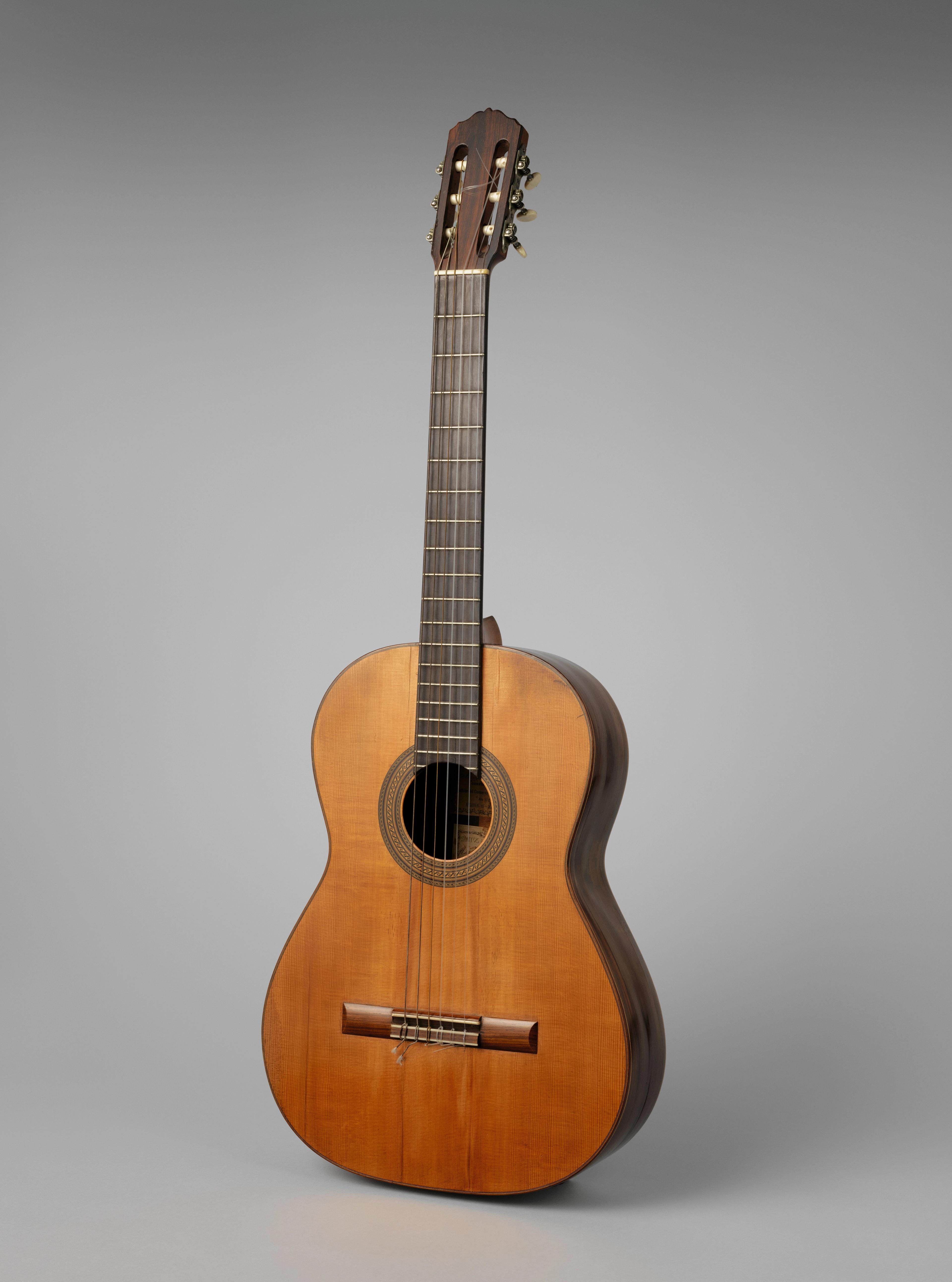 Vintage wooden acoustic guitar stands upright against a gray background, showcasing its warm brown body, strings, fretboard, and sound hole.
